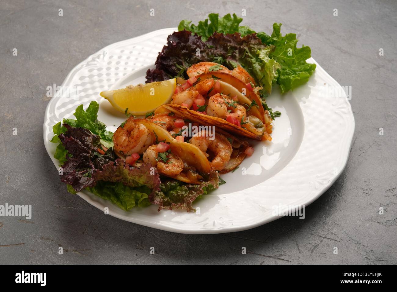 Tacos aux crevettes avec laitue fraîche et coin de citron servis sur une assiette blanche sur une surface en pierre grise Banque D'Images