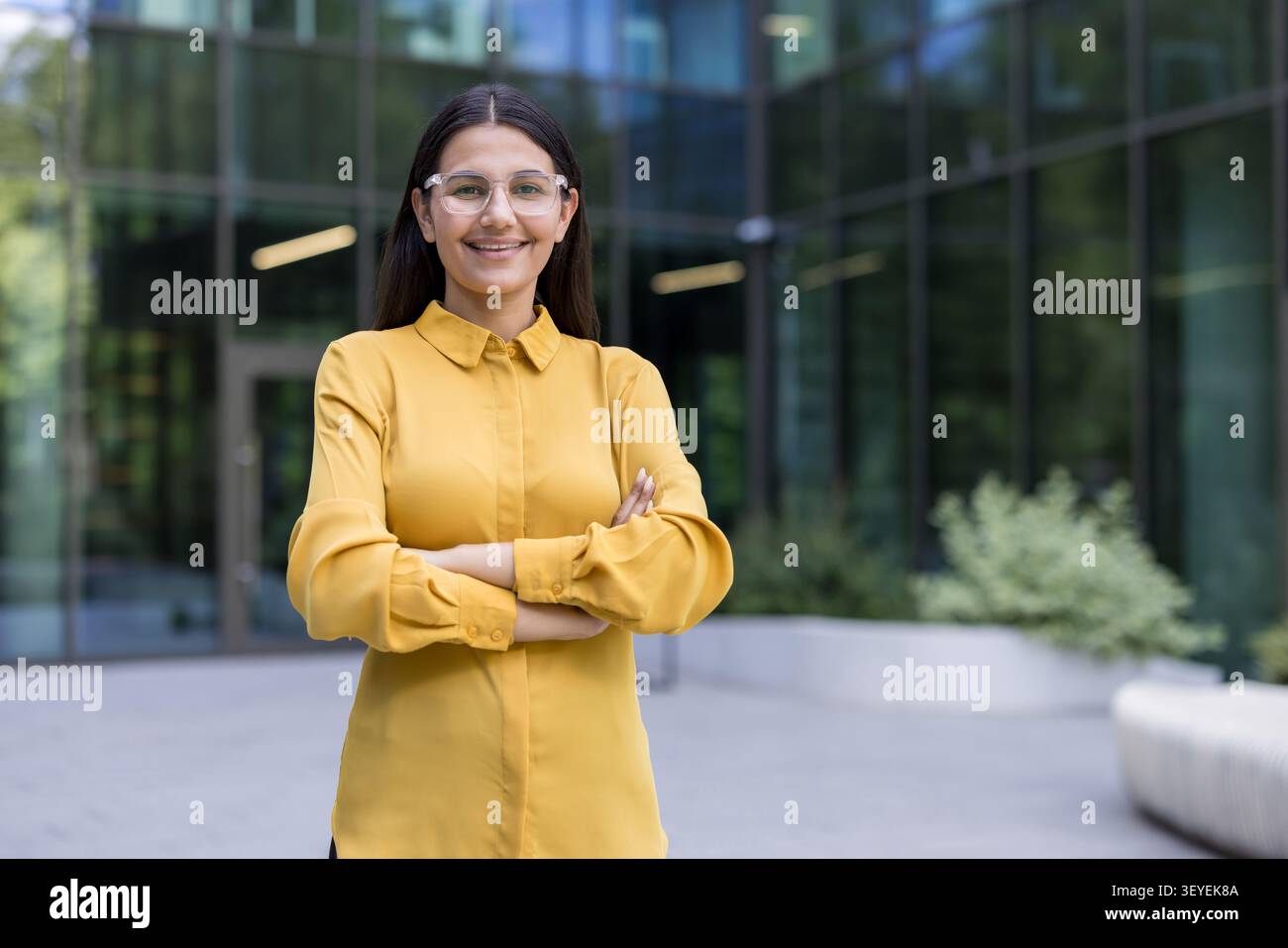 Femme d'affaires confiante portant des lunettes et une chemise jaune, debout à l'extérieur avec les bras croisés sur fond de bâtiment d'entreprise moderne, reflétant le succès et la croissance professionnelle Banque D'Images