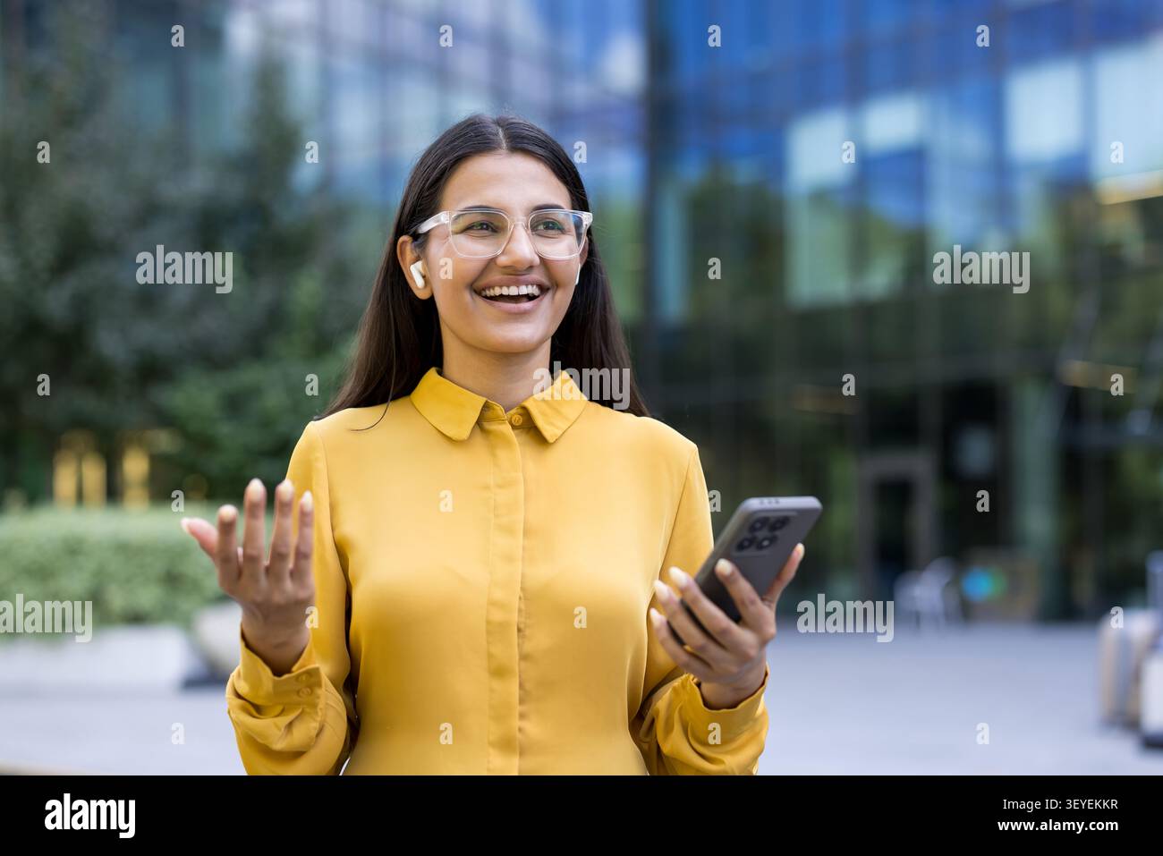 Femme en lunettes et chemise jaune souriant joyeusement tout en interagissant sur un smartphone. Port d'écouteurs sans fil. Debout à l'extérieur d'un bâtiment moderne Banque D'Images