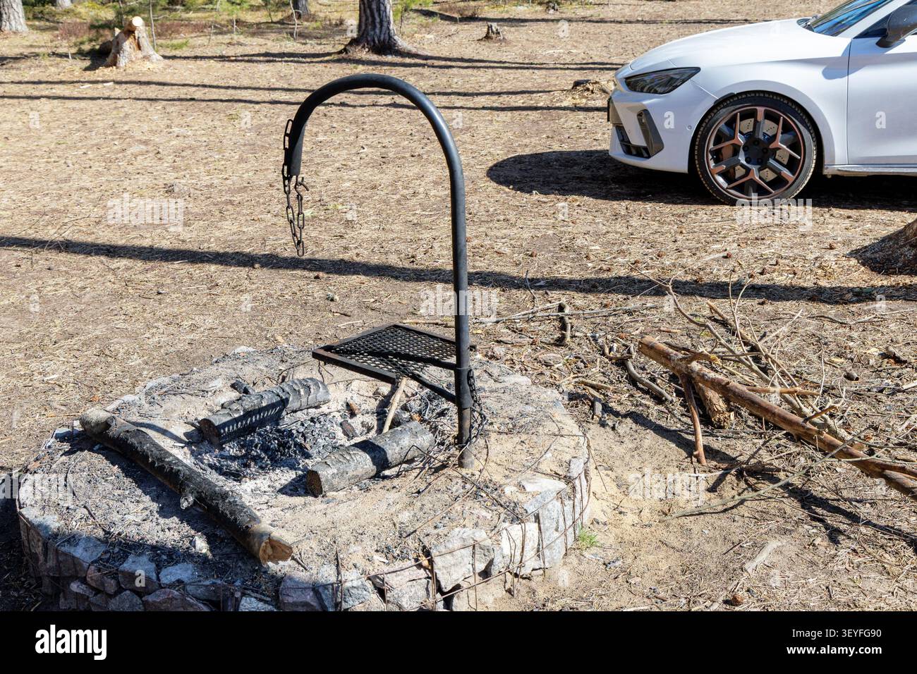 Une photo en grand angle d'un feu de camp en pierre vide avec une structure de grille métallique, avec une voiture moderne blanche garée à proximité dans une forêt. Banque D'Images