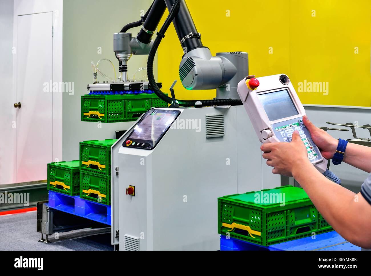 Ingénieur industriel opérant un bras robotisé automatisé avec un contrôleur à pendentif pédagogique pour soulever des caisses vertes dans un entrepôt d'usine moderne Banque D'Images