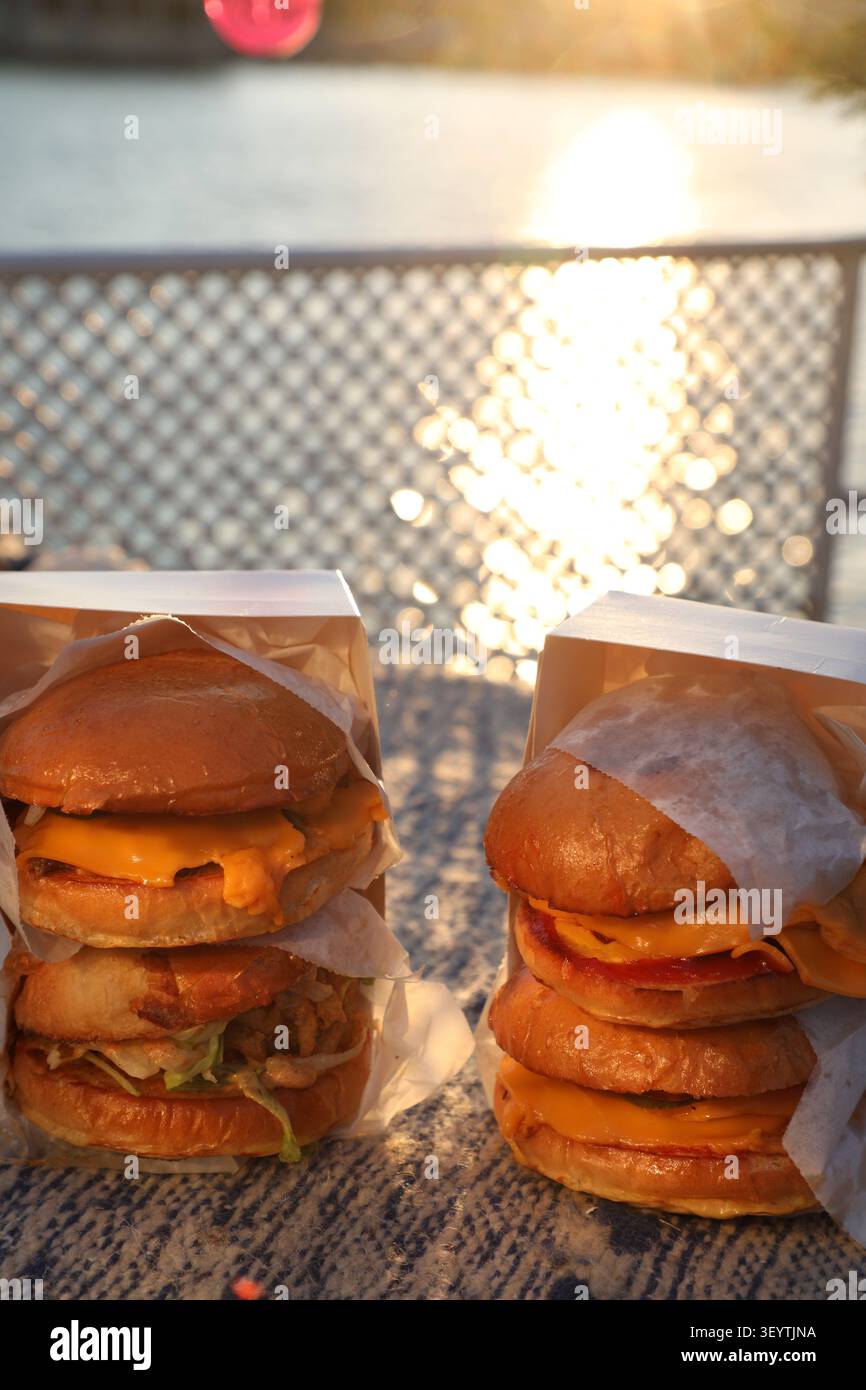 Deux hamburgers sont posés sur une table près de l'eau. Le soleil se couche à l'horizon et illumine les hamburgers. Les emballages sont en papier et s Banque D'Images