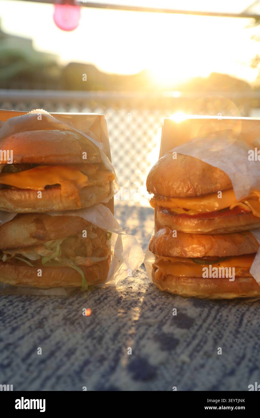 Deux hamburgers sont placés sur une table à l'extérieur. Le soleil se couche à l'horizon, illuminant les burgers. Ils sont emballés dans du papier. L' Banque D'Images