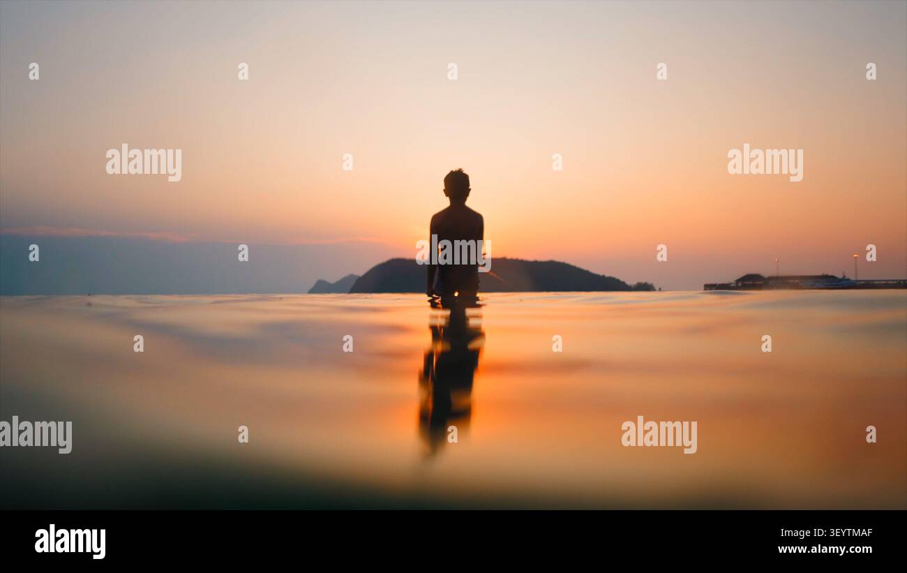 Enfant solitaire debout dans l'eau paisible regardant le coucher de soleil doré à l'horizon Banque D'Images