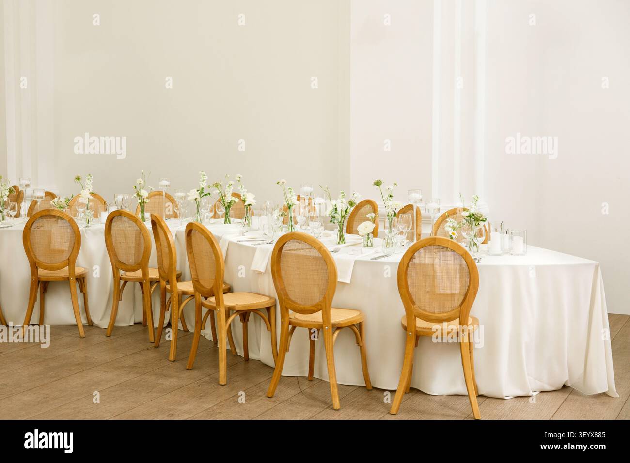 Une longue table de réception de mariage est dressée avec des nappes blanches, des chaises en rotin et des vases floraux délicats dans un cadre intérieur minimaliste Banque D'Images
