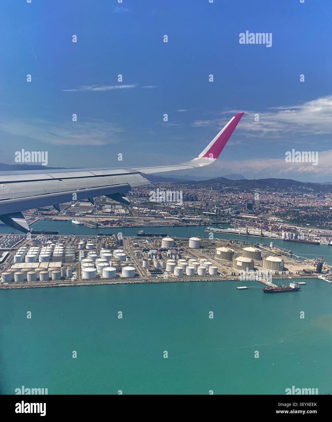 Vue aérienne panoramique capturée depuis une fenêtre d'avion montrant l'aile au-dessus d'un port industriel avec des réservoirs de stockage de pétrole, des cargos et une turquoise Banque D'Images
