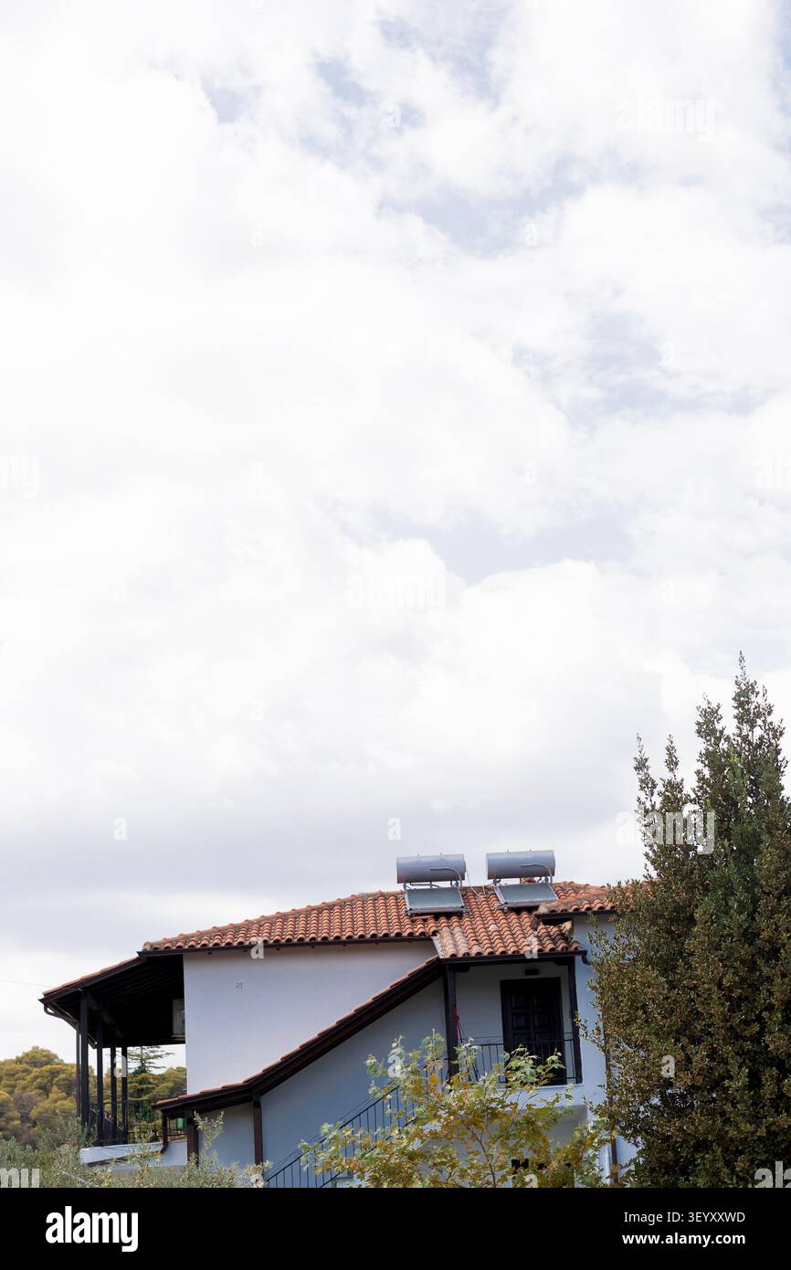Une maison blanche de deux étages avec un toit de tuiles en terre cuite et des chauffe-eau solaires. Le bâtiment dispose d'un balcon et de grandes fenêtres entourées de verdure Banque D'Images