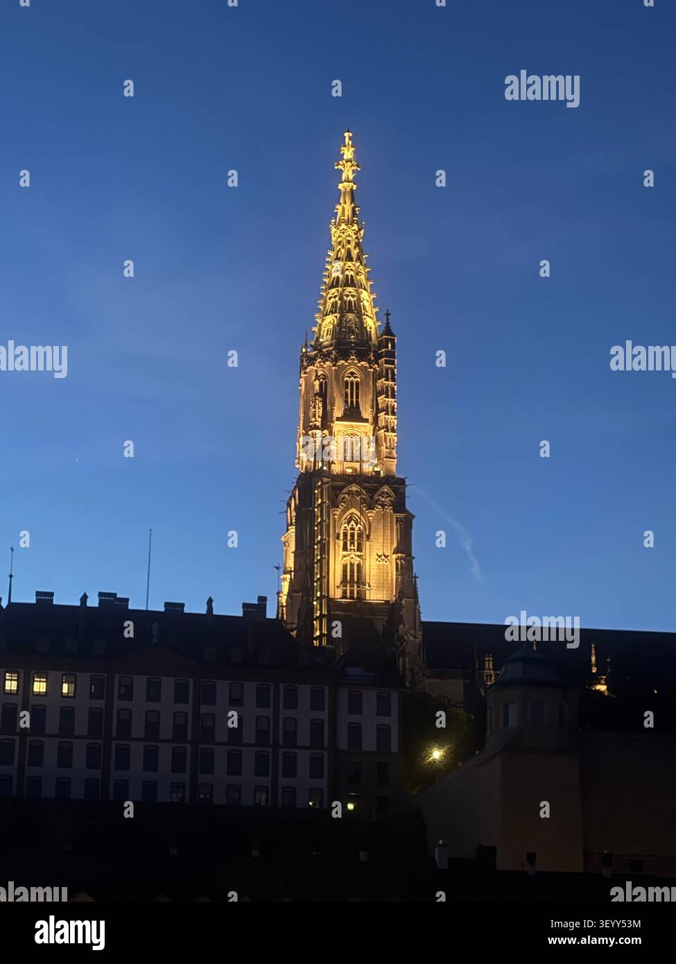 La tour gothique illuminée de la cathédrale de Berne dominant les toits de la ville contre un ciel crépusculaire bleu profond, Berne, Suisse - Image de stock capturée avec un smartphone