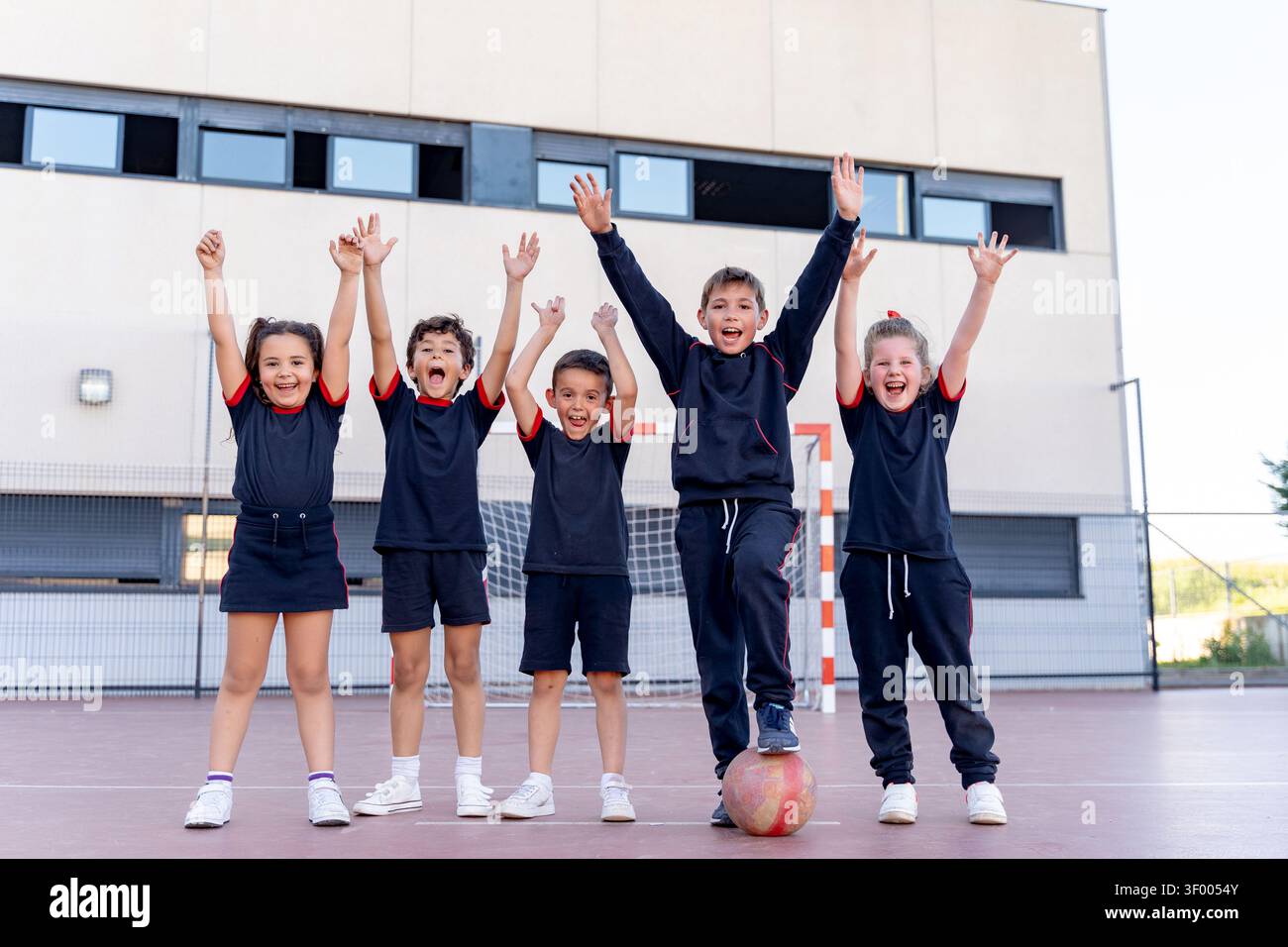 Groupe d'enfants excités en uniforme acclamant avec les bras levés, debout sur un terrain de sport après avoir joué un match Banque D'Images