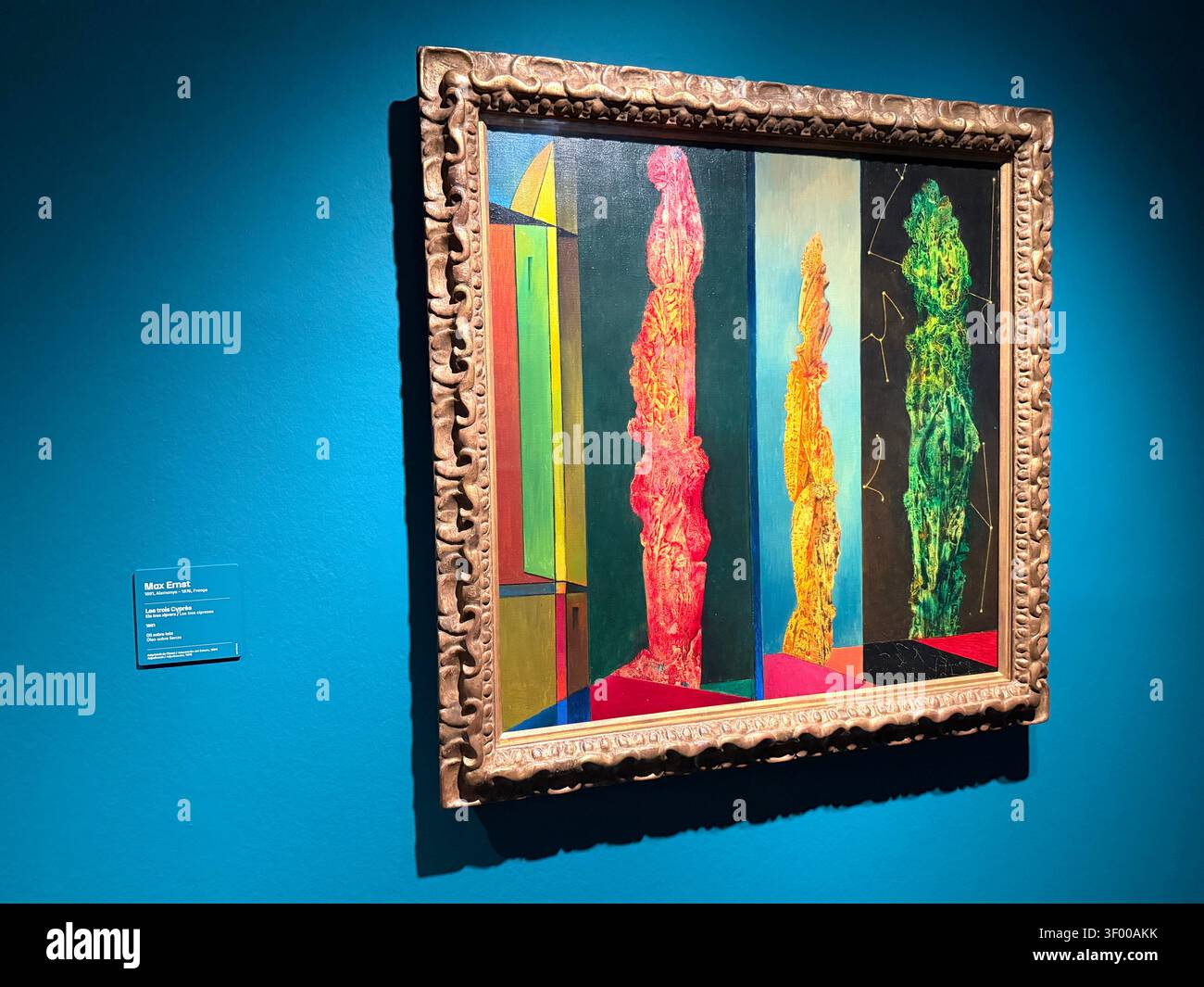 Peinture à l'huile les trois cyprès de Max Ernst créée en 1951 dans un cadre doré sur un mur bleu - Image de stock capturée avec un smartphone