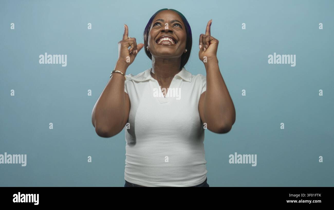 Femme en polo blanc pointe les index vers le haut et sourit largement montrant les dents en studio avec fond turquoise ; célébration de joie. Banque D'Images