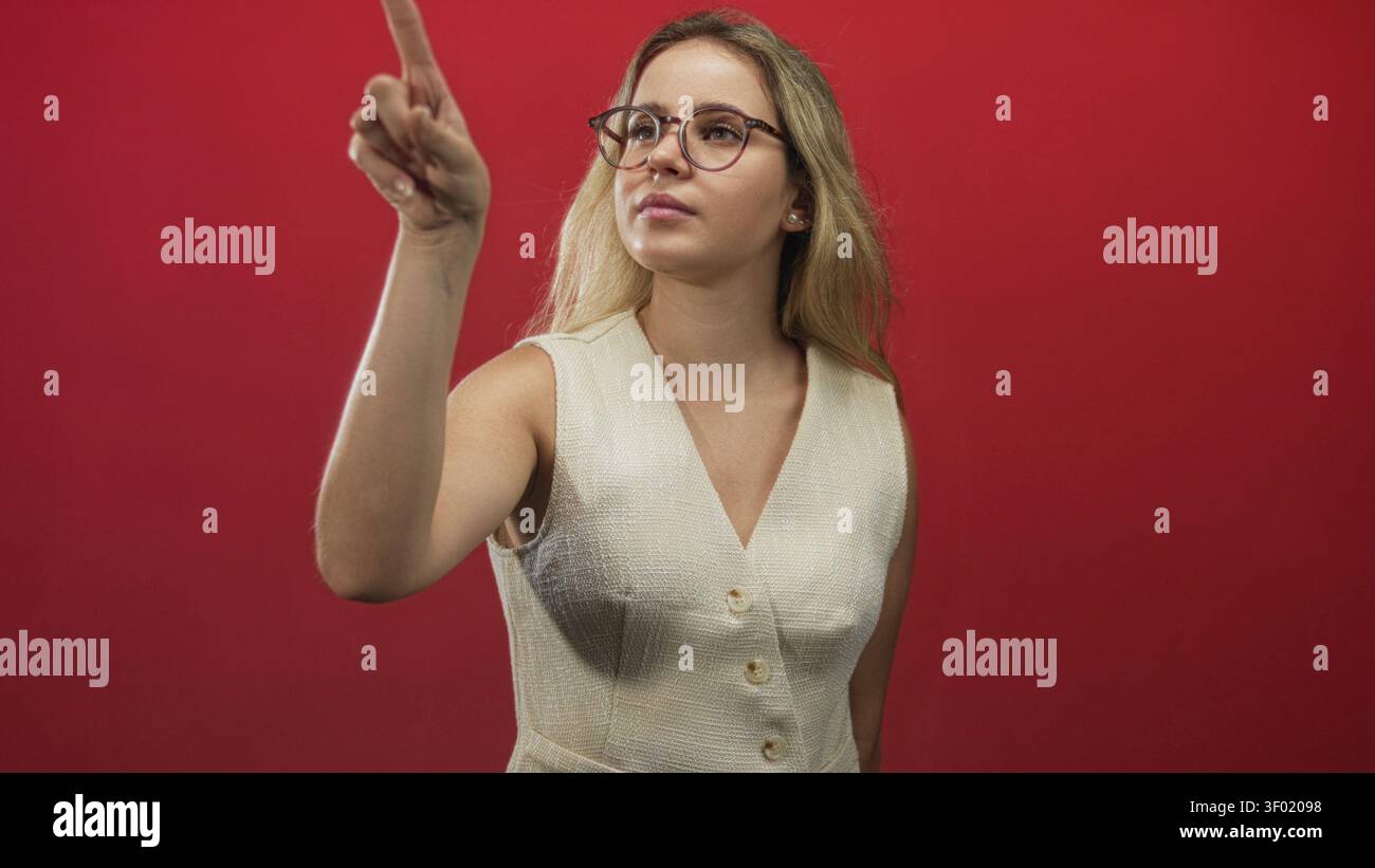 Jeune femme blonde dans des lunettes pointe doigt vers le haut dans le studio contre le mur rouge tout en portant un gilet crème sans manches et regardant vers l'avant ; mise au point de confiance. Banque D'Images