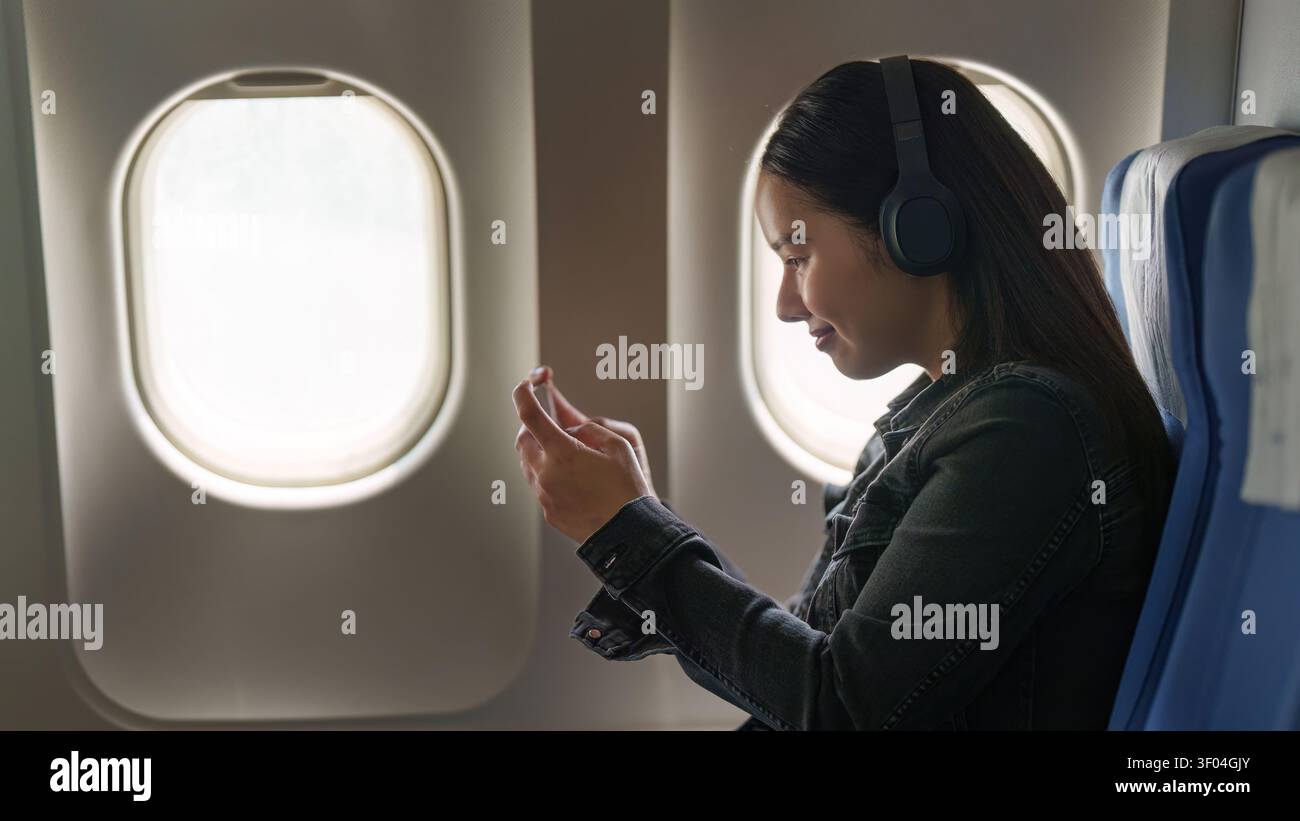 Femme asiatique avec casque utilisant un smartphone dans un avion pour se divertir pendant le voyage Banque D'Images