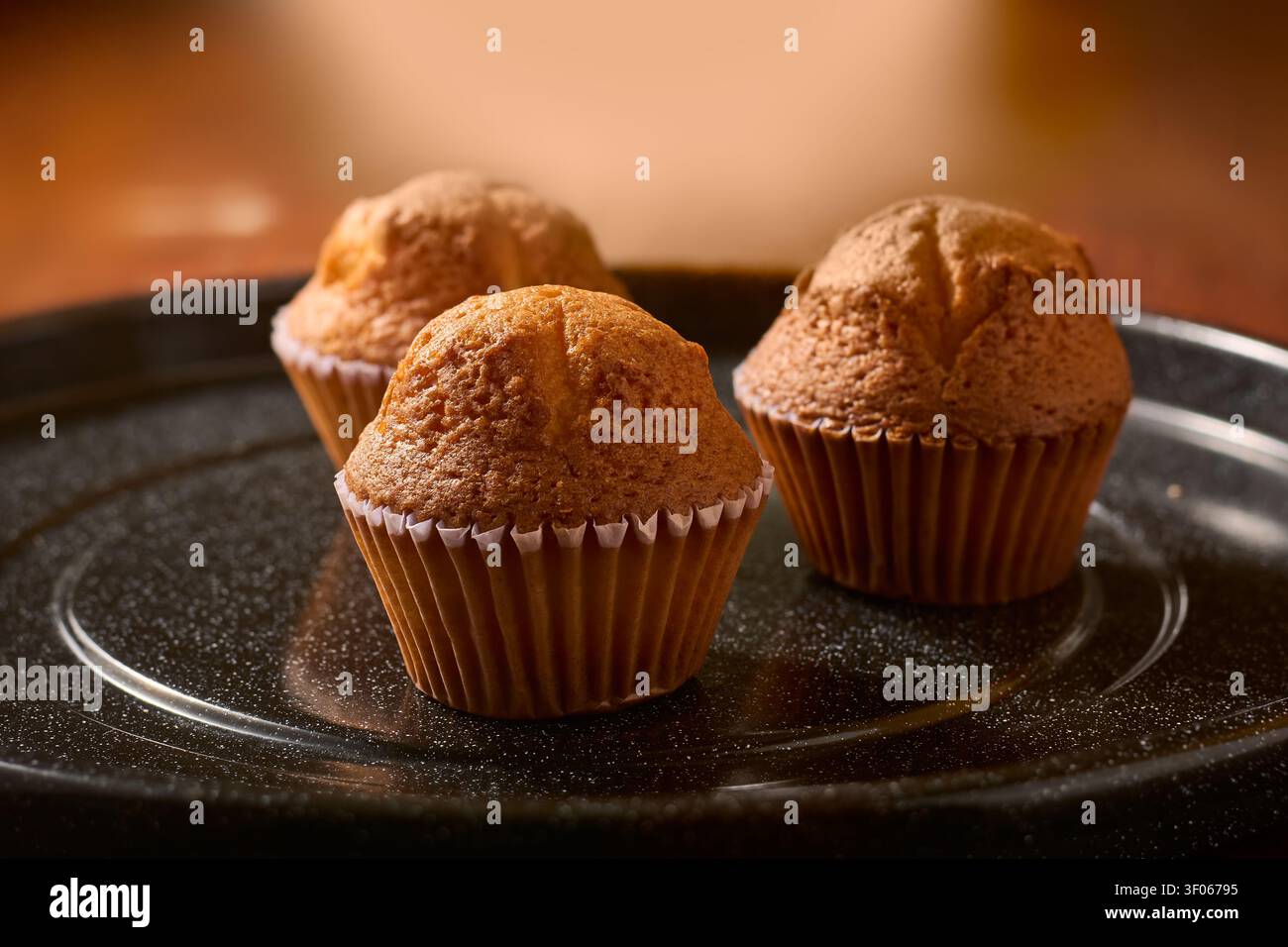 trois muffins fraîchement cuits sur une assiette noire texturée, gâteaux moelleux dorés dans des moules en papier, vue de côté avec mise au point douce et espace de copie, chaud fait maison Banque D'Images