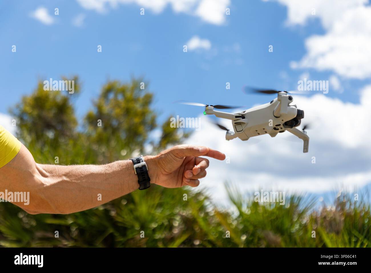 Gros plan de la main d'une personne atteignant un petit drone quadcopter planant à l'extérieur par une journée brillante, avec un ciel bleu, des nuages et des arbres verts dans le Banque D'Images