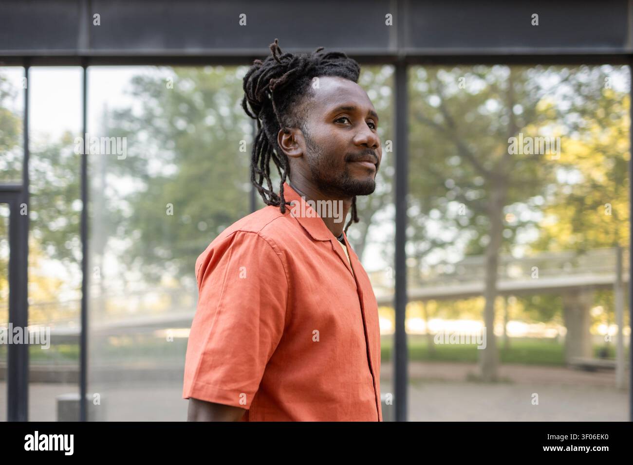Portrait profil de côté d'un homme cubain avec des dreadlocks dans une chemise orange, debout à l'extérieur près d'un bâtiment en verre moderne, regardant devant dans un naturel doux Banque D'Images
