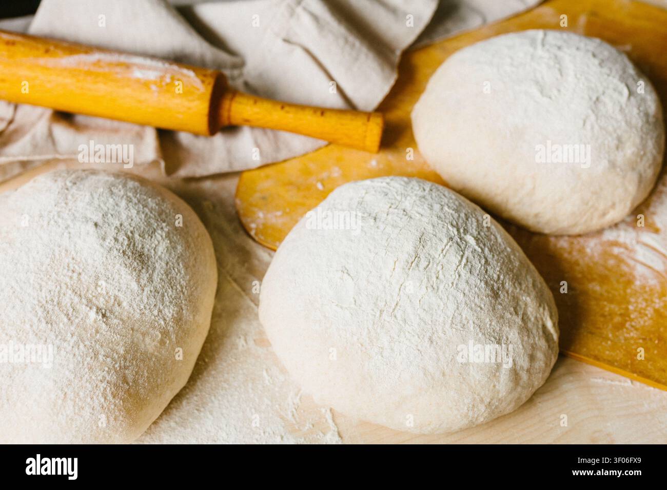Trois boules de pâte farinées reposent sur une planche rustique à côté d'un rouleau à pâtisserie, prêtes pour la fermentation et la cuisson. Préparation artisanale authentique de pain utilisant ou Banque D'Images