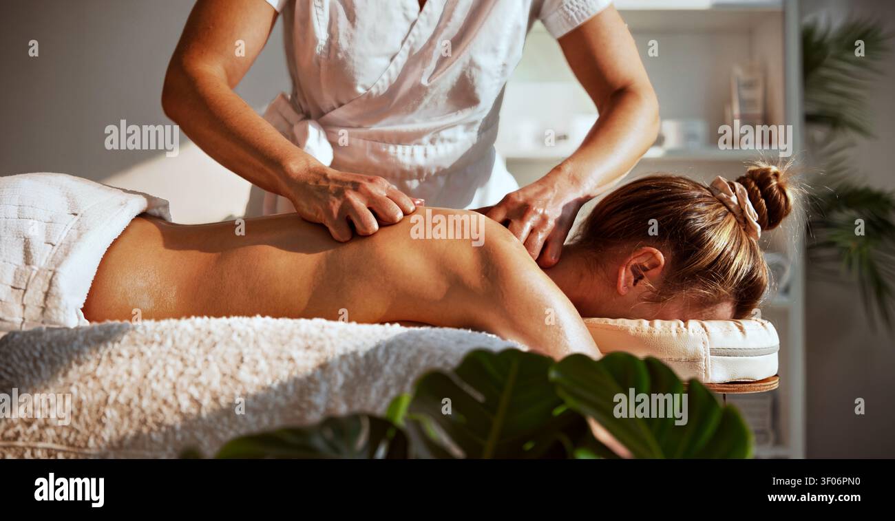 Thérapeute de spa bien-être biophile donne un massage en profondeur des tissus du dos à une femme sur une table recouverte d'une serviette blanche, à côté de feuillages luxuriants de Monstera tropicale Banque D'Images