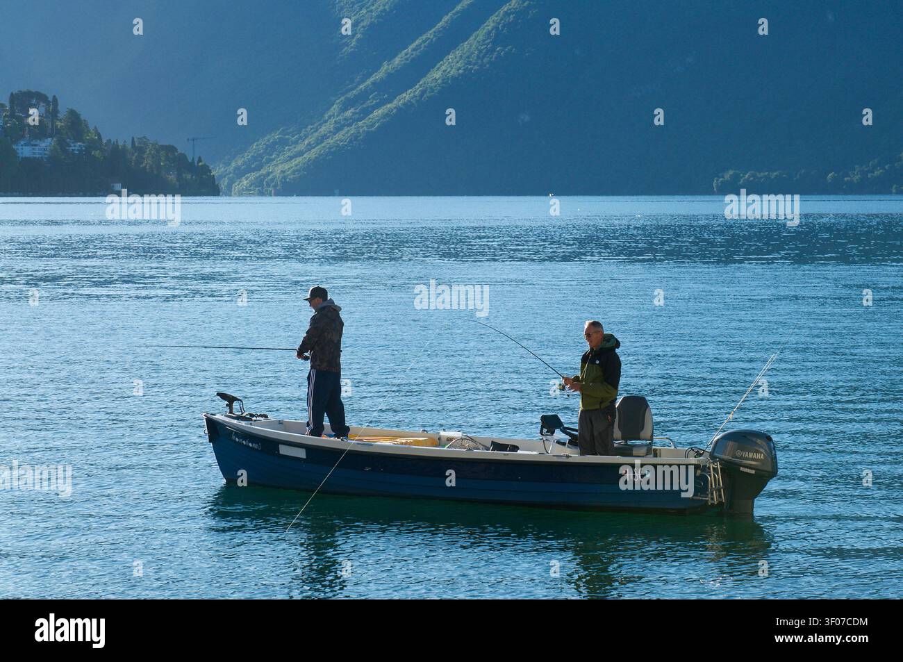 Lugano, Tessin, Suisse – 5 juin 2026 : Vue panoramique de deux pêcheurs avec des cannes à pêche debout sur un petit bateau à moteur au petit matin. Banque D'Images