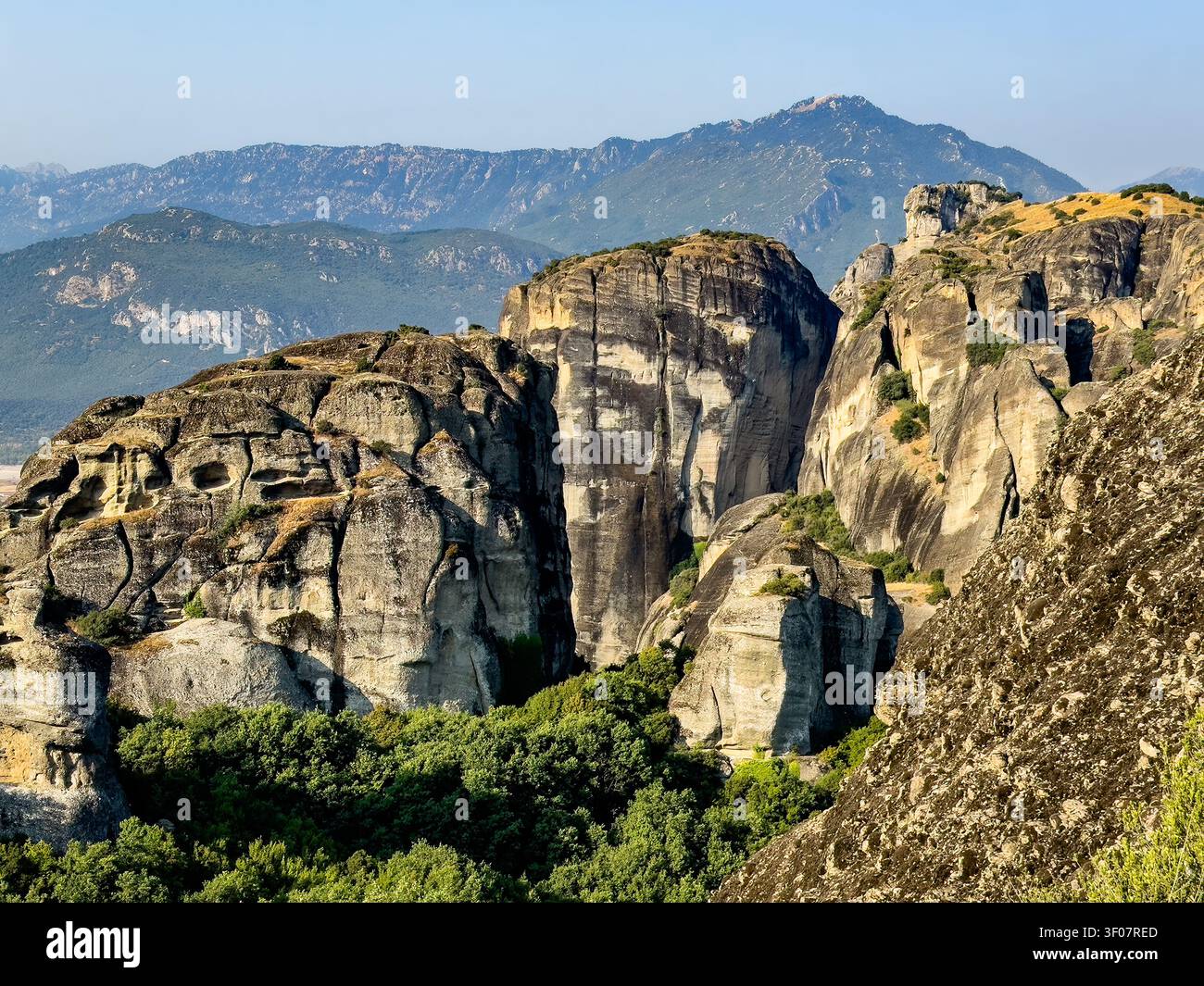 Kalambaka, Grèce – 24 juillet 2024 : groupe de monastères Meteora. Un groupe de rochers escarpés avec des grottes. Feuillage vert au premier plan. Ciel bleu. Chaîne de montagnes o Banque D'Images