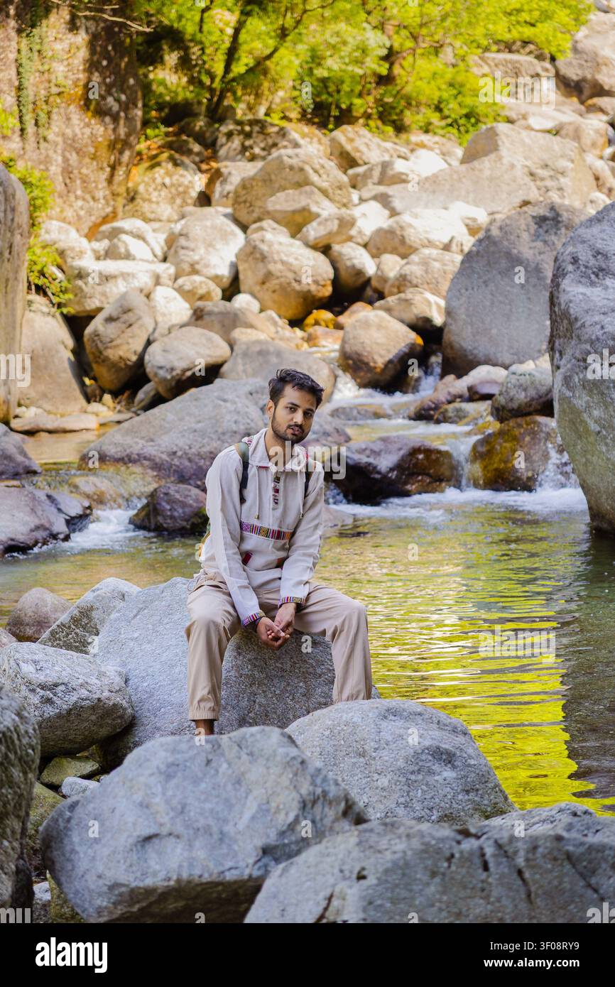 Un jeune homme avec un sac à dos est assis sur un gros rocher à côté d'un ruisseau de montagne qui coule dans un paysage rocheux. Banque D'Images