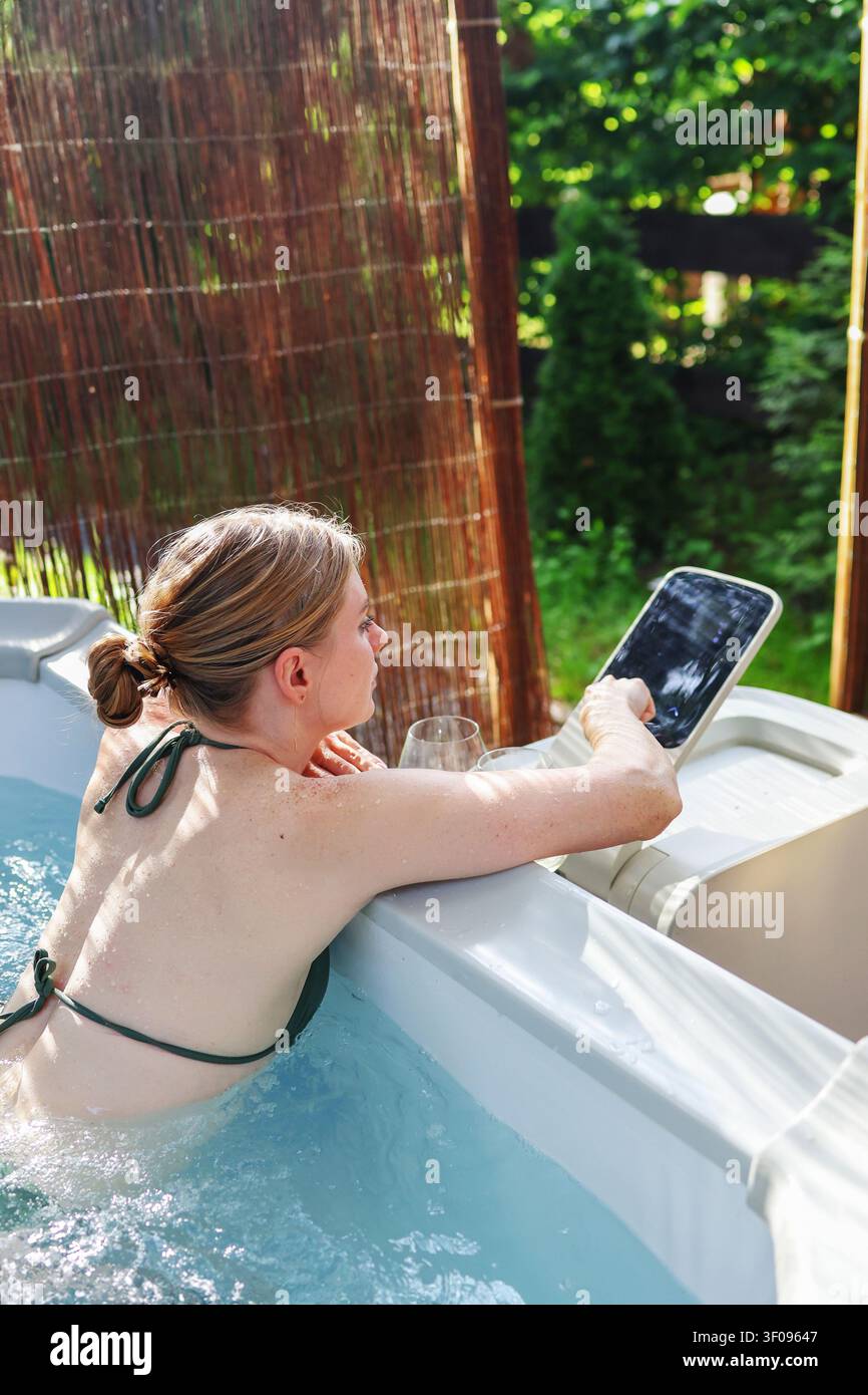 Jeune femme se détendant dans un jacuzzi de luxe avec panneau tactile dans l’arrière-cour, créant une expérience spa et de soins personnels paisible. Banque D'Images