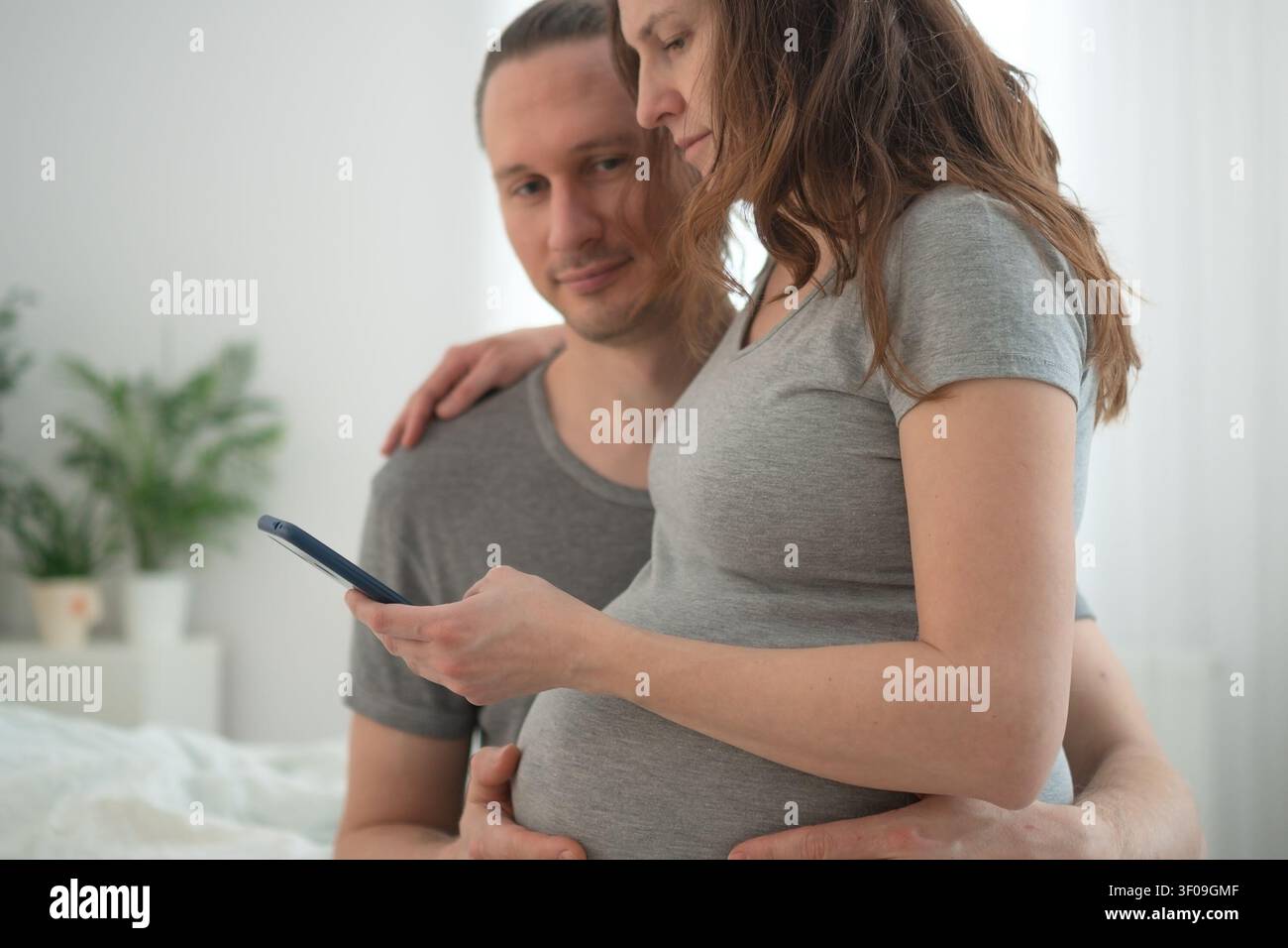 Femme enceinte et son mari debout ensemble dans une chambre lumineuse partageant les nouvelles de la grossesse lors d'un appel téléphonique. La mère enceinte a le bras autour de la taille tandis que son partenaire tient le téléphone et communique avec la famille. Banque D'Images