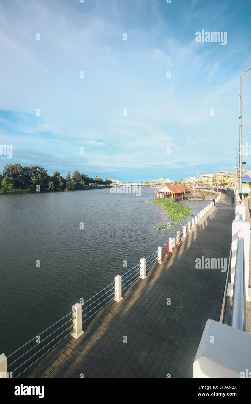 Vue grand angle de la promenade pavée du front de mer de Mae Klong s'incurvant le long de l'eau avec un pavillon traditionnel à Ratchaburi, Thaïlande. Banque D'Images