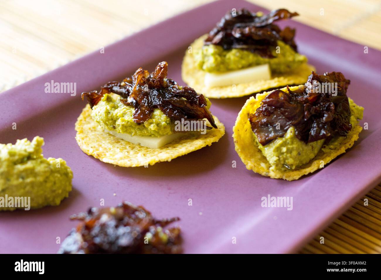 Trois canapés de chips de maïs avec crème d'asperges, fromage et radicchio caramélisé sur une assiette rectangulaire violette posée sur un set de table en bambou Banque D'Images