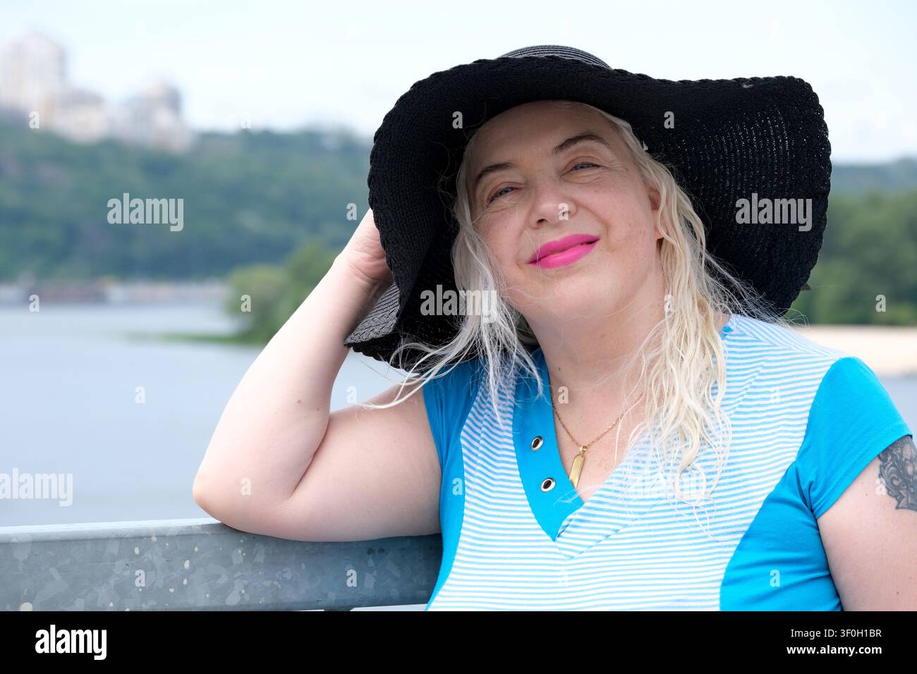 Belle femme mûre souriante de plus de 50 ans portant un chapeau à larges bords noir et une tenue d'été bleue, posant en plein air au bord de l'eau par une chaude journée ensoleillée. Nat Banque D'Images