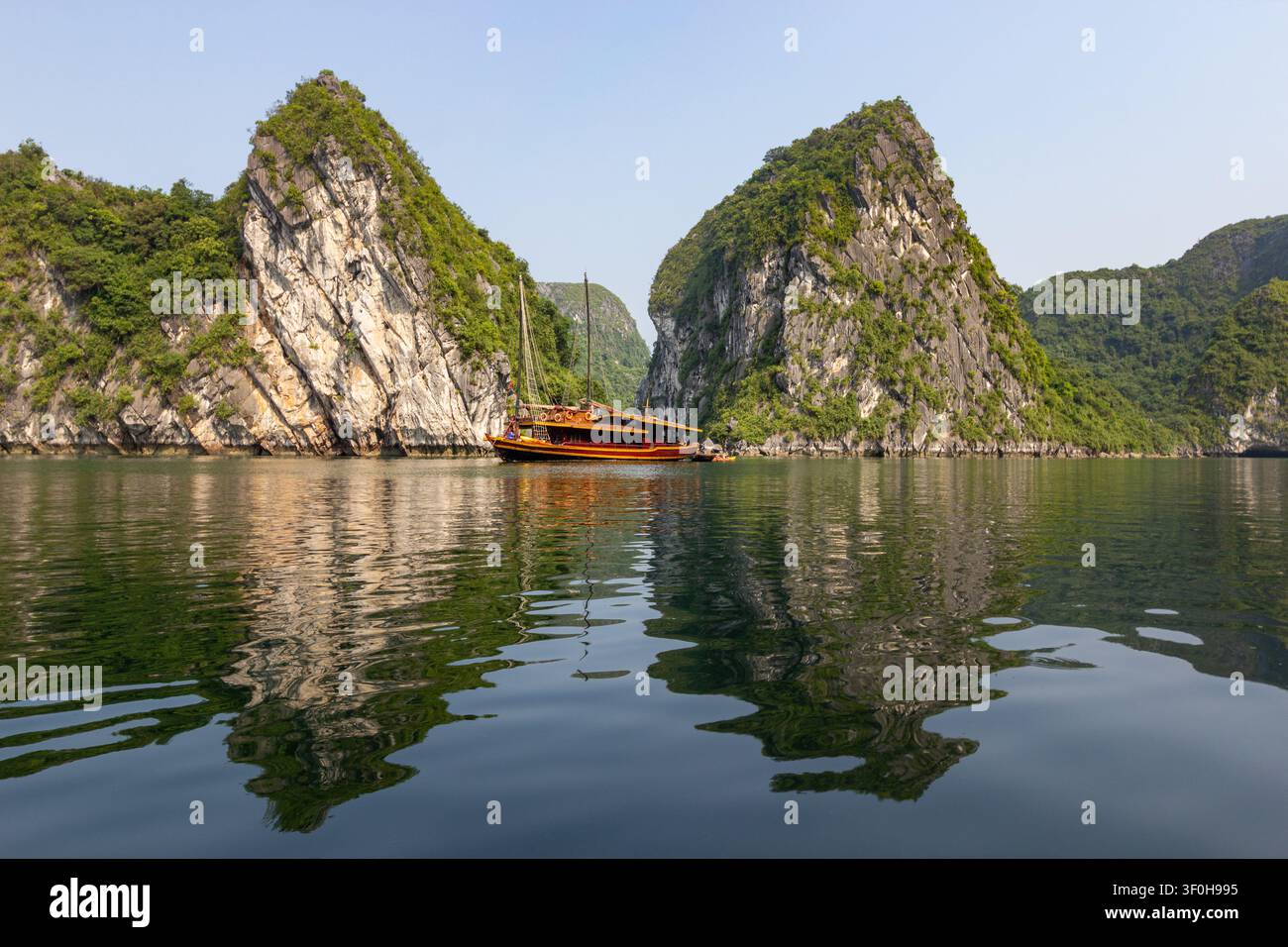 Grandes formations karstiques en calcaire et un bateau-junk touristique traditionnel en bois réfléchissant dans la baie d'Halong, Vietnam Banque D'Images