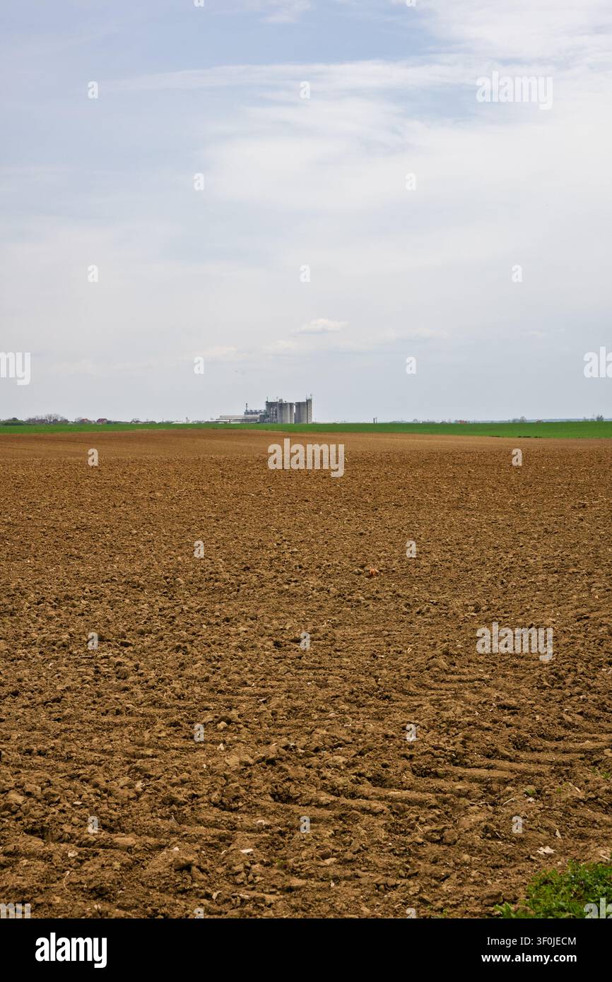 Un vaste champ labouré s'étend jusqu'à l'horizon où des silos à grains distants s'élèvent sous un vaste ciel nuageux, évoquant l'agriculture rurale, l'espace ouvert et le calme Banque D'Images