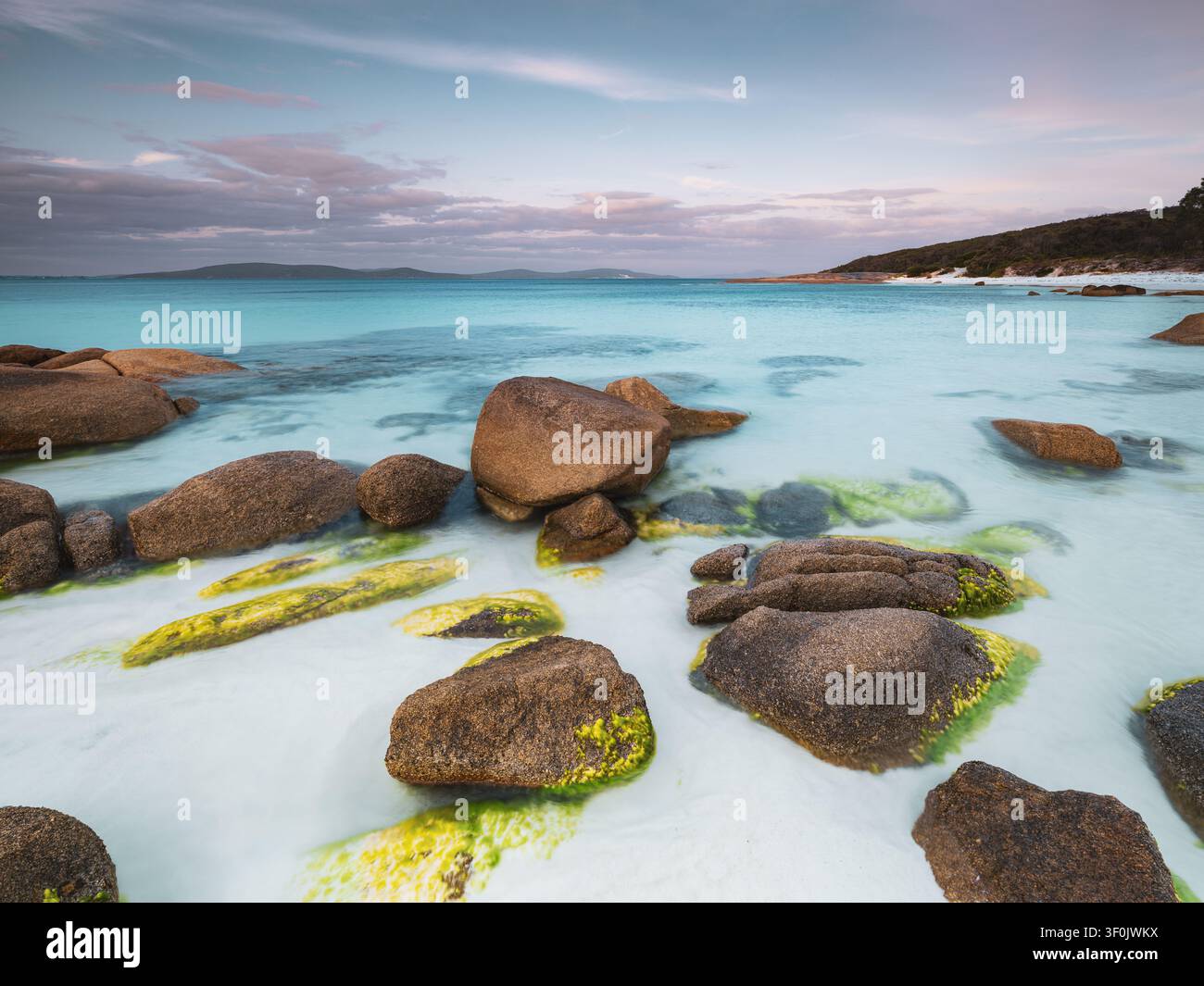 Vue de Whalers Cove avec de gros rochers de granit couverts de mousse reposant dans une eau turquoise peu profonde sous un ciel pastel doux à Albany, WA, Australie. Banque D'Images