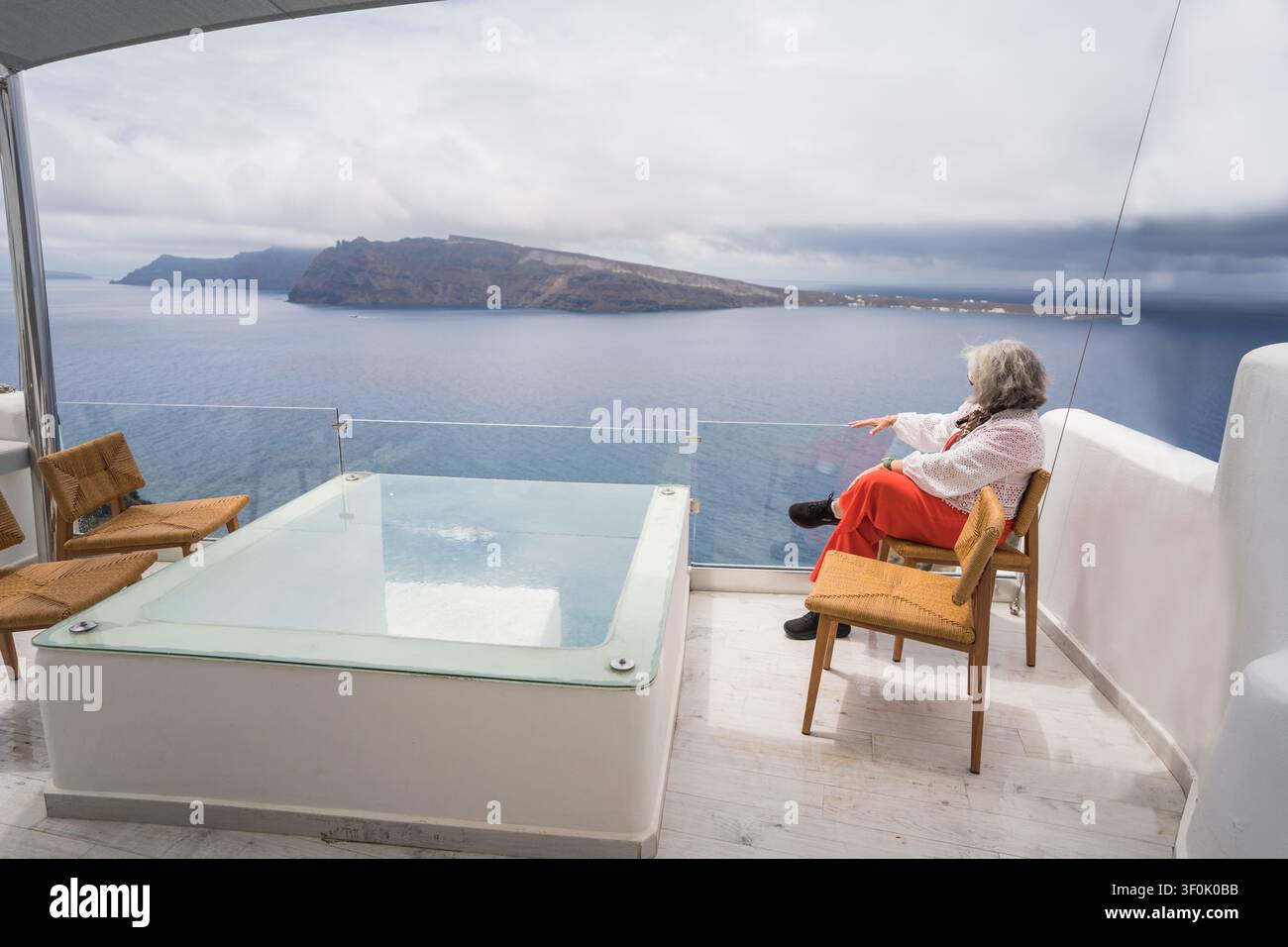 Sénior assise sur une terrasse d'hôtel de luxe à Santorin, en Grèce, profitant d'une vue calme sur la mer par-dessus la caldera avec un jacuzzi privé, des chaises modernes, un Banque D'Images
