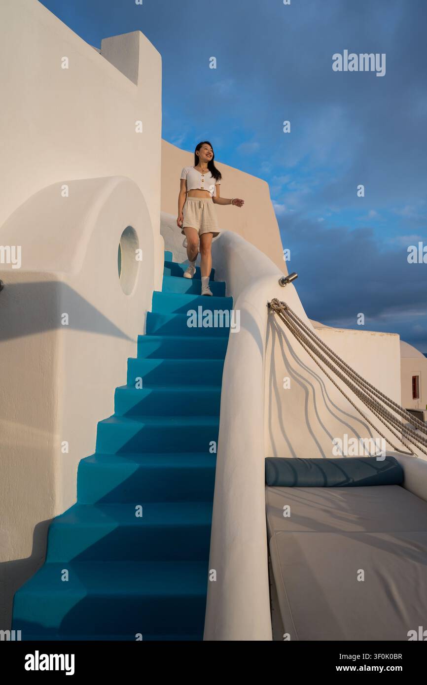 Femme asiatique montant des escaliers bleu vif dans un hôtel de luxe à Santorini, en Grèce, entourée d'architecture blanchée à la chaux et d'une lumière chaude de coucher de soleil sous un Banque D'Images