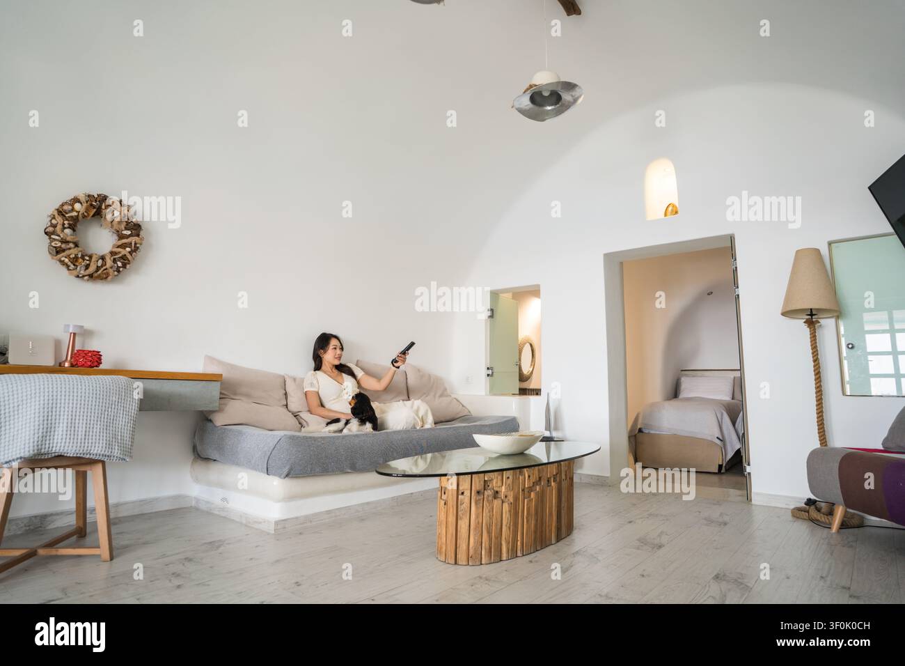 Femme asiatique se détendant sur un canapé dans une suite d'hôtel de luxe à Santorin, en Grèce, tenant un smartphone dans une pièce blanche lumineuse de style grotte avec une décoration minimaliste Banque D'Images