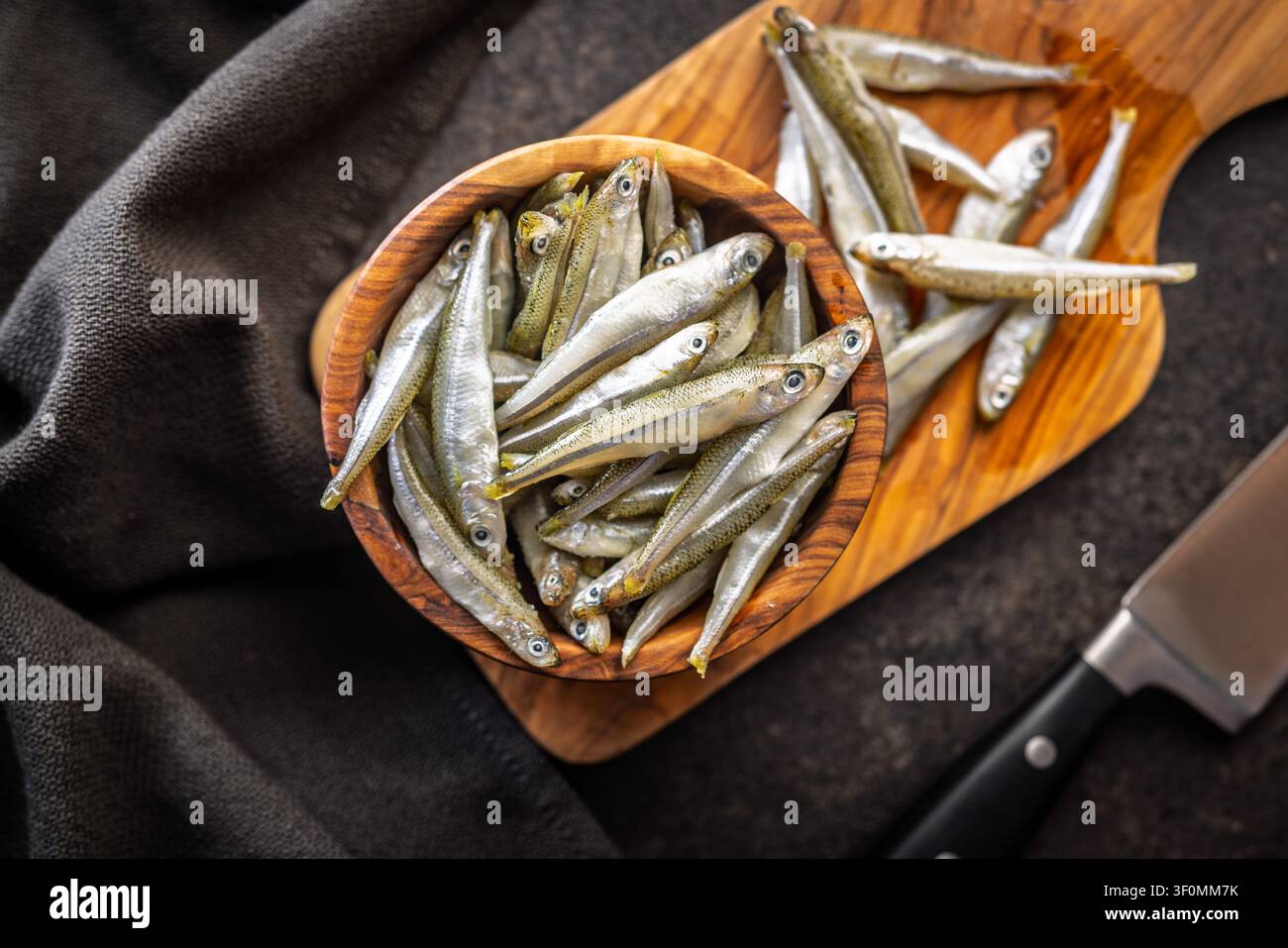 Petits poissons frais empilés dans un bol en bois sur une surface rustique, idéaux pour les recettes de fruits de mer, les entrées ou les plats méditerranéens. La texture naturelle et la présentation simple mettent en valeur la fraîcheur et l'utilisation culinaire. Banque D'Images