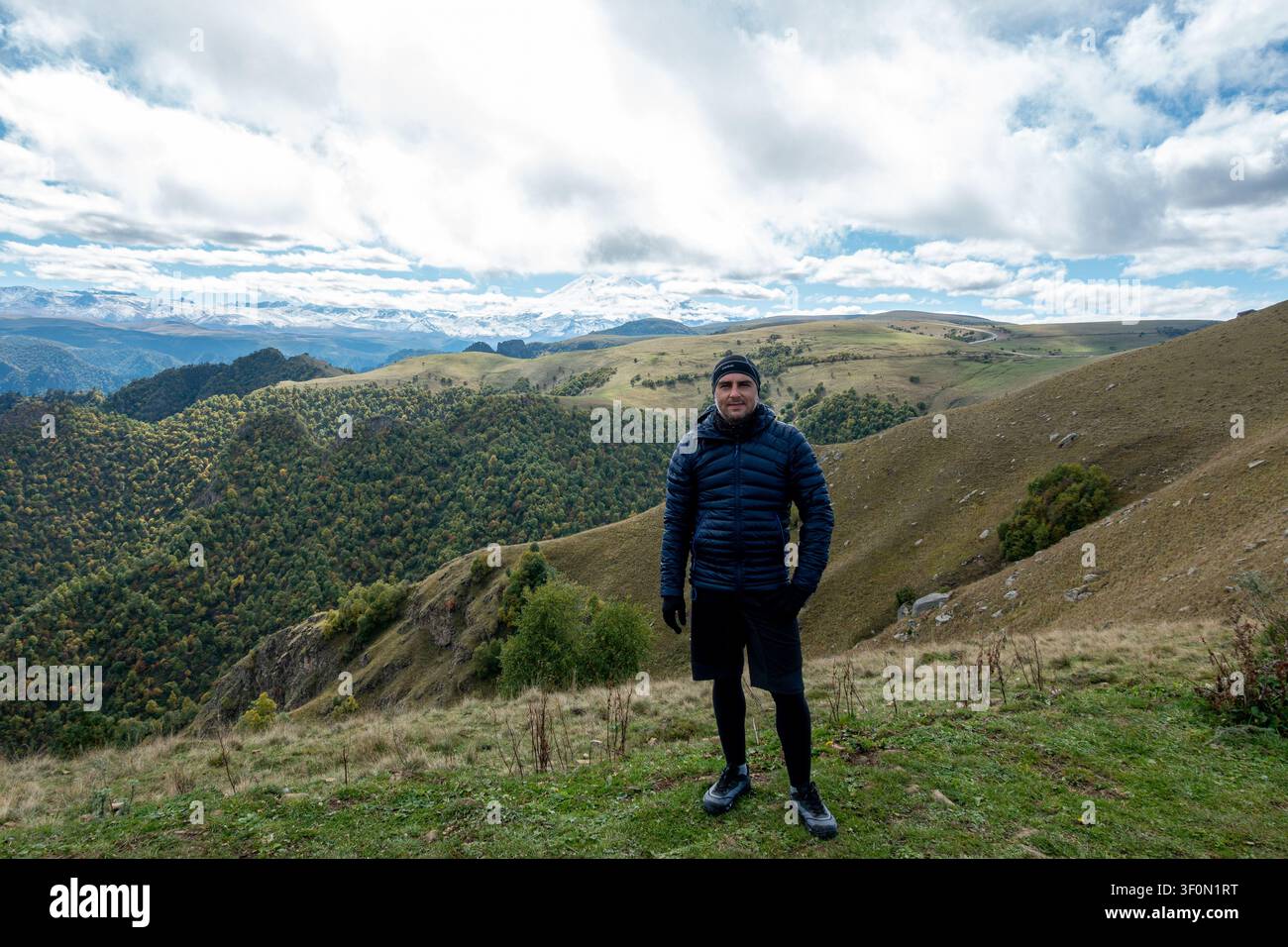 Un randonneur vêtu d'une doudoune bleu marine et d'un bonnet se tient sur une crête de prairie alpine dans le Caucase. Banque D'Images