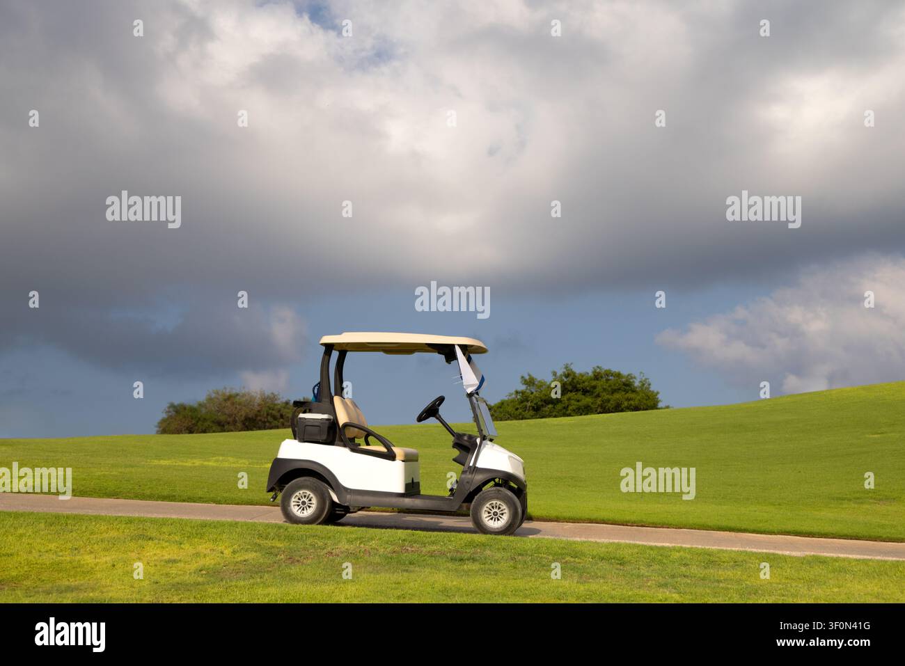 Chariot de golf blanc garé sur un chemin pavé d'un magnifique terrain de golf avec des collines vertes ondulantes et des nuages d'orage dramatiques dans le ciel. Image de stock commerciale parfaite pour les concepts de loisirs et de sports. Banque D'Images