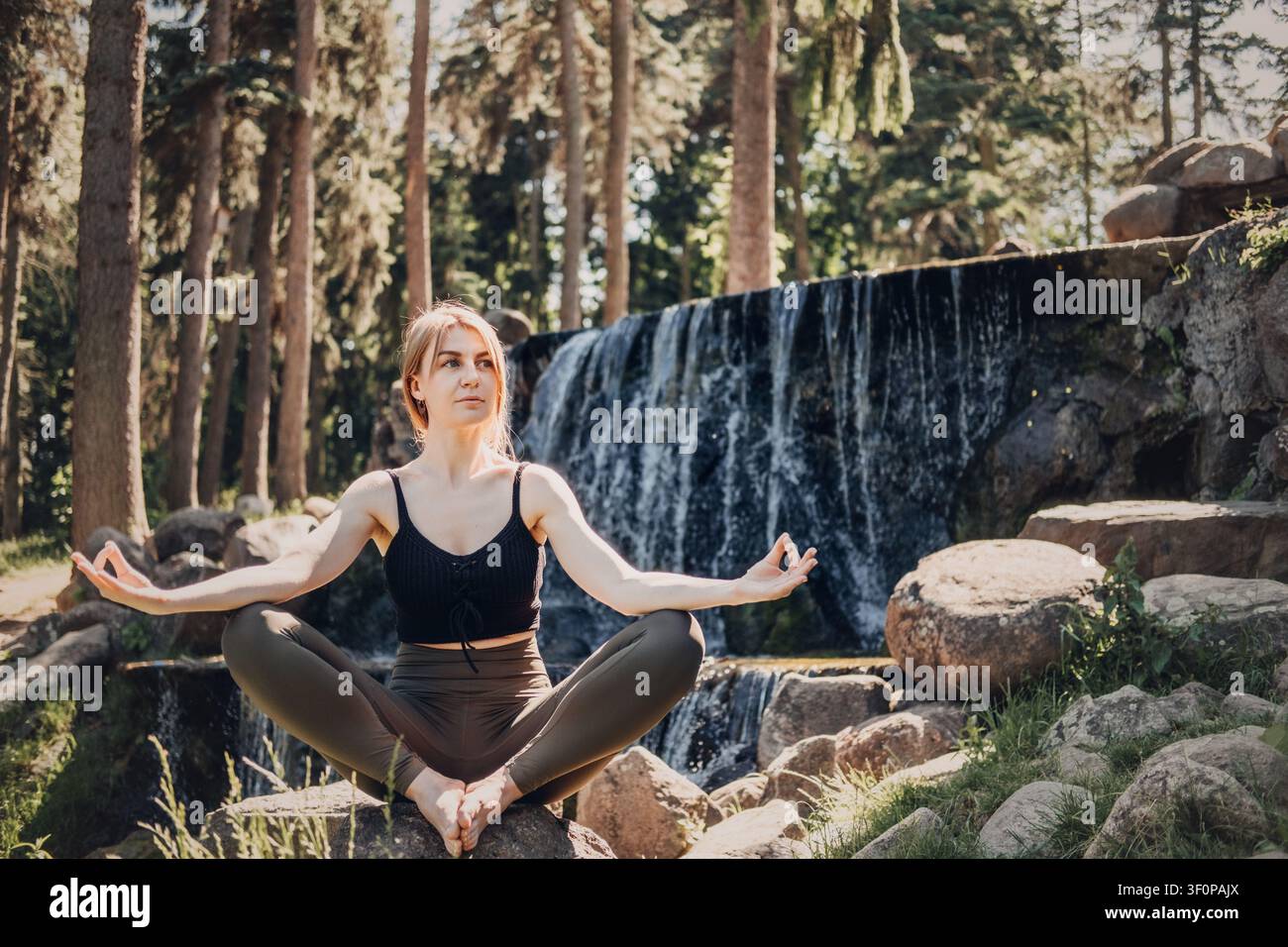 Jeune femme assise en posture de yoga en plein air près d'une cascade. Femme pratiquant le yoga en plein air dans une clairière forestière. Retreat de détox numérique, santé mentale Banque D'Images