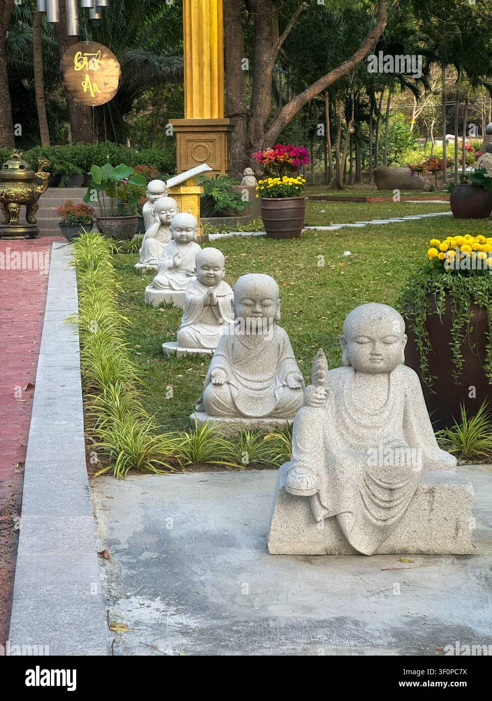 Range de petites statues de moines en pierre dans un jardin de temple paisible. Adorables sculptures de bouddha novices bordent une pelouse verdoyante. Fleurs colorées en pots, t - Image de stock capturée avec un smartphone