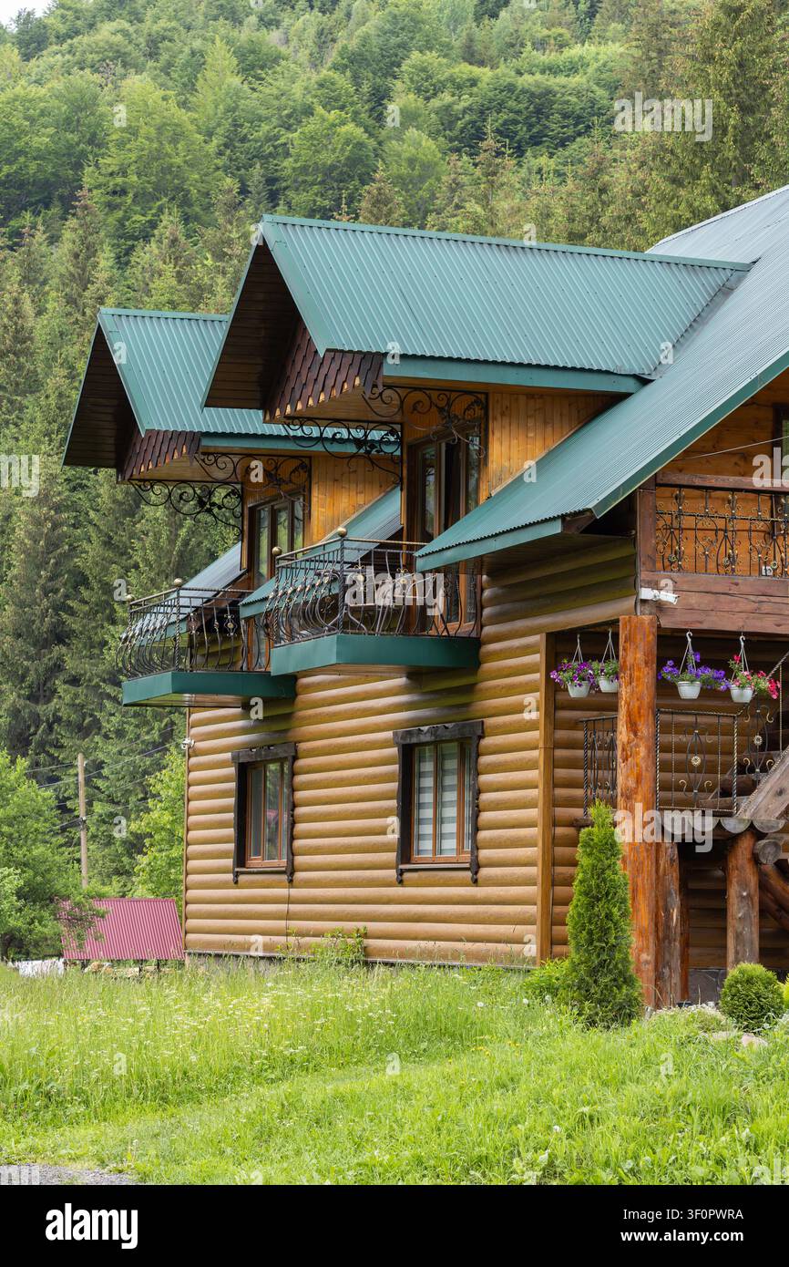 Un chalet en rondins à plusieurs étages avec des balcons en fer se dresse contre une pente abrupte couverte d’une forêt dense de conifères et de feuillus. Banque D'Images