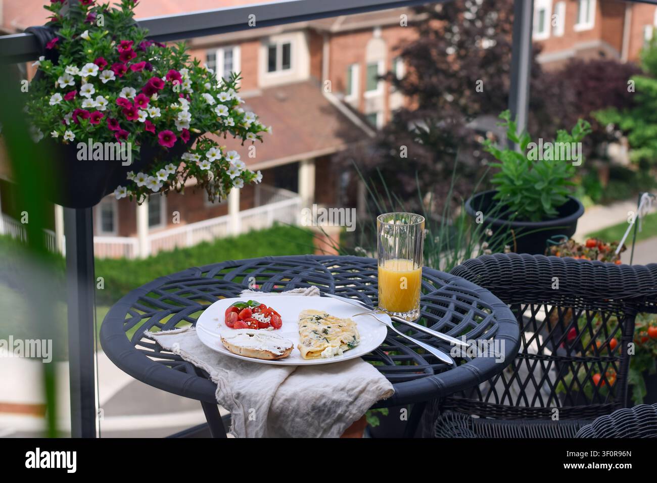 Plan large au niveau des yeux montrant une assiette de petit-déjeuner avec omelette et jus d'orange sur une table en métal noir avec un panier de fleurs suspendu et une maison en briques floue en arrière-plan Banque D'Images