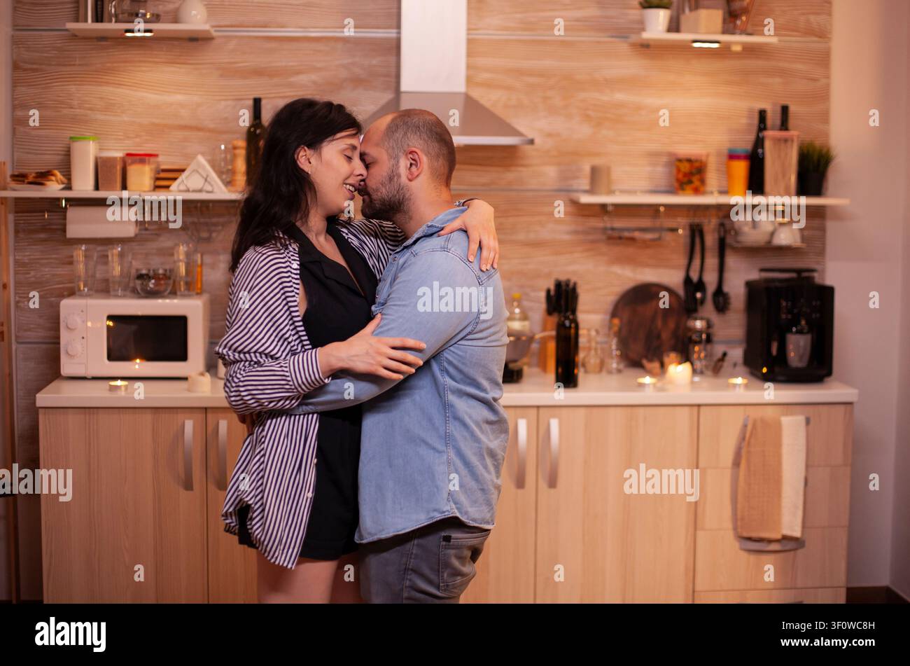 Le mari serre sa femme dans ses bras lors d'une célébration de leur relation à la maison alors que le couple sourit, partage un dîner et profite d'un moment romantique chaleureux dans la cuisine, reliant câlin de couple et dîner romantique. Banque D'Images