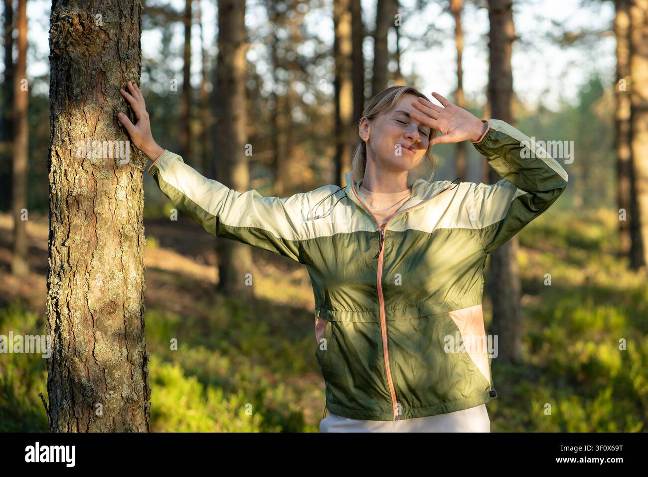 Hiker joyeuse profitant d'une promenade en forêt et ressentant des émotions positives, debout à côté d'un arbre en souriant. Banque D'Images