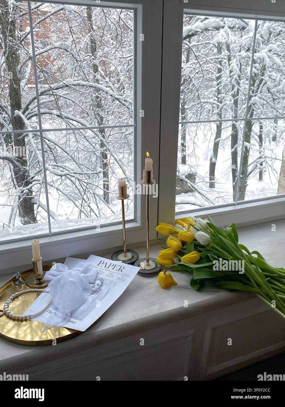 Moscou, Russie : 5 mars 2022 - Des tulipes fraîches jaunes et blanches disposées sur un rebord de fenêtre avec des bougies allumées surplombant un paysage hivernal enneigé - Image de stock capturée avec un smartphone