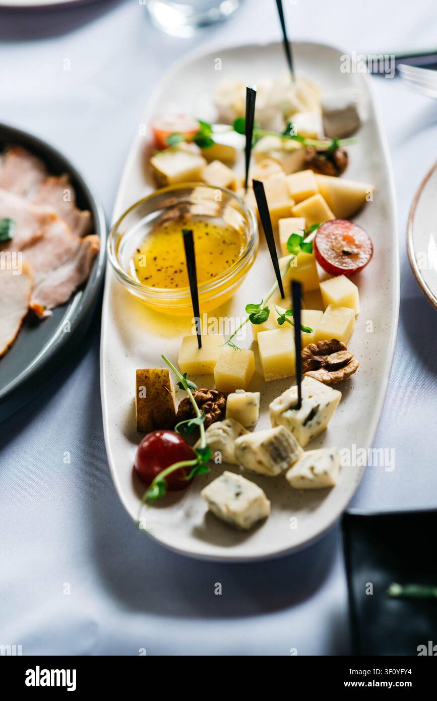 Planche à fromage élégante mettant en vedette une variété de fromages à pâte dure et bleus accompagnés de miel, de raisins rouges, de noix, de micro-pousses fraîches et de pics à cocktail sur un plat en céramique blanche. Apéritif raffiné magnifiquement présenté sur une table de restaurant. Banque D'Images