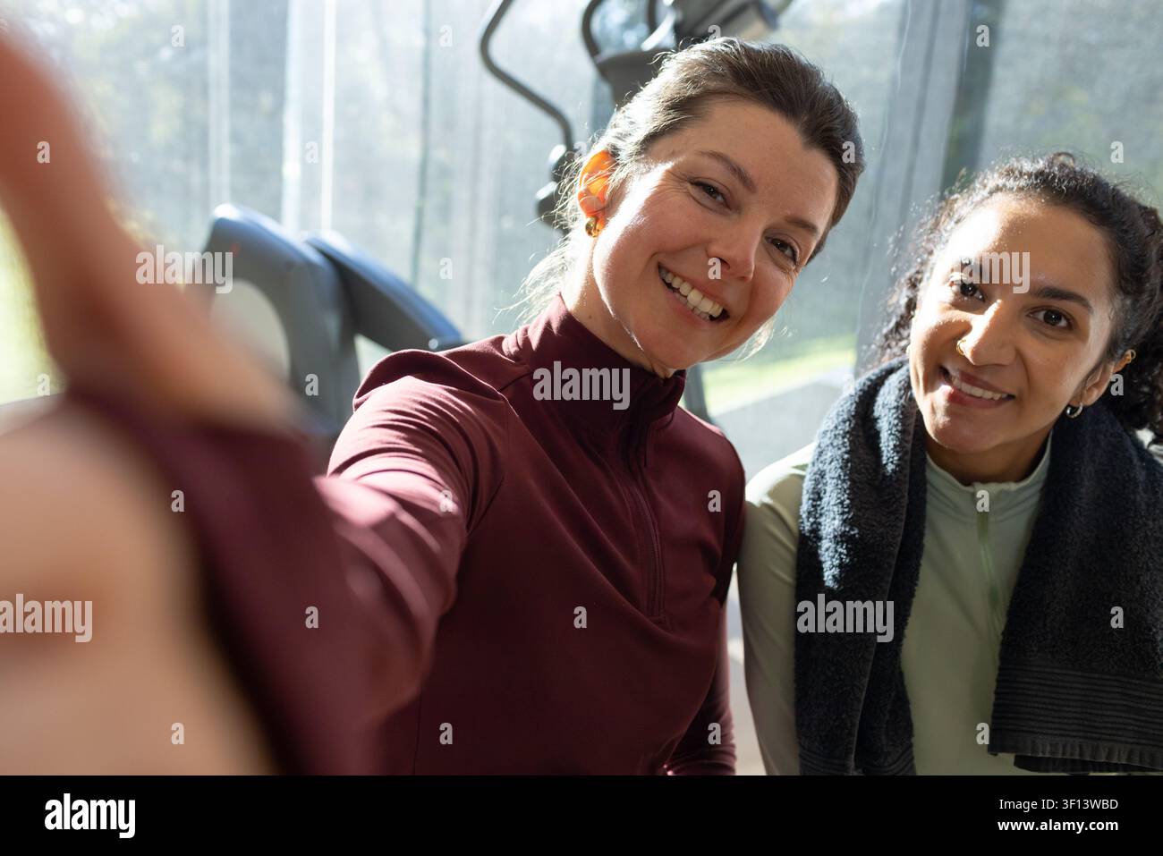 Deux amies souriantes et posant pour un selfie dans une salle de sport, portant des hauts vert marron près des tapis de course Banque D'Images