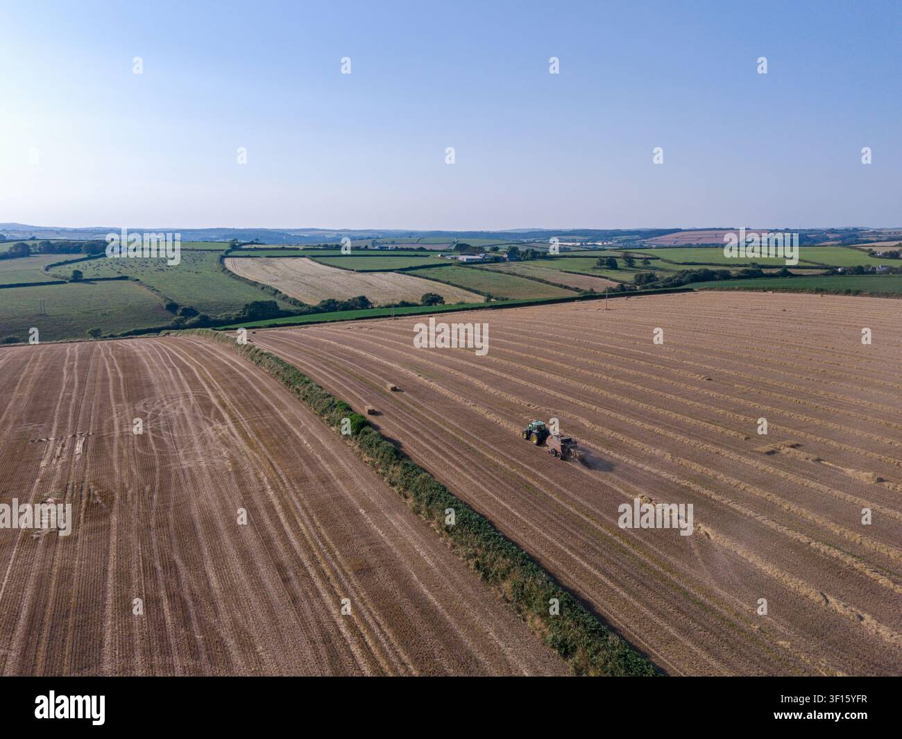 Vue aérienne d'un tracteur faisant des balles de foin dans un grand champ récolté lors d'une soirée d'été. Les collines ondulantes et les champs de Cornwall sont visibles en arrière-plan. Banque D'Images