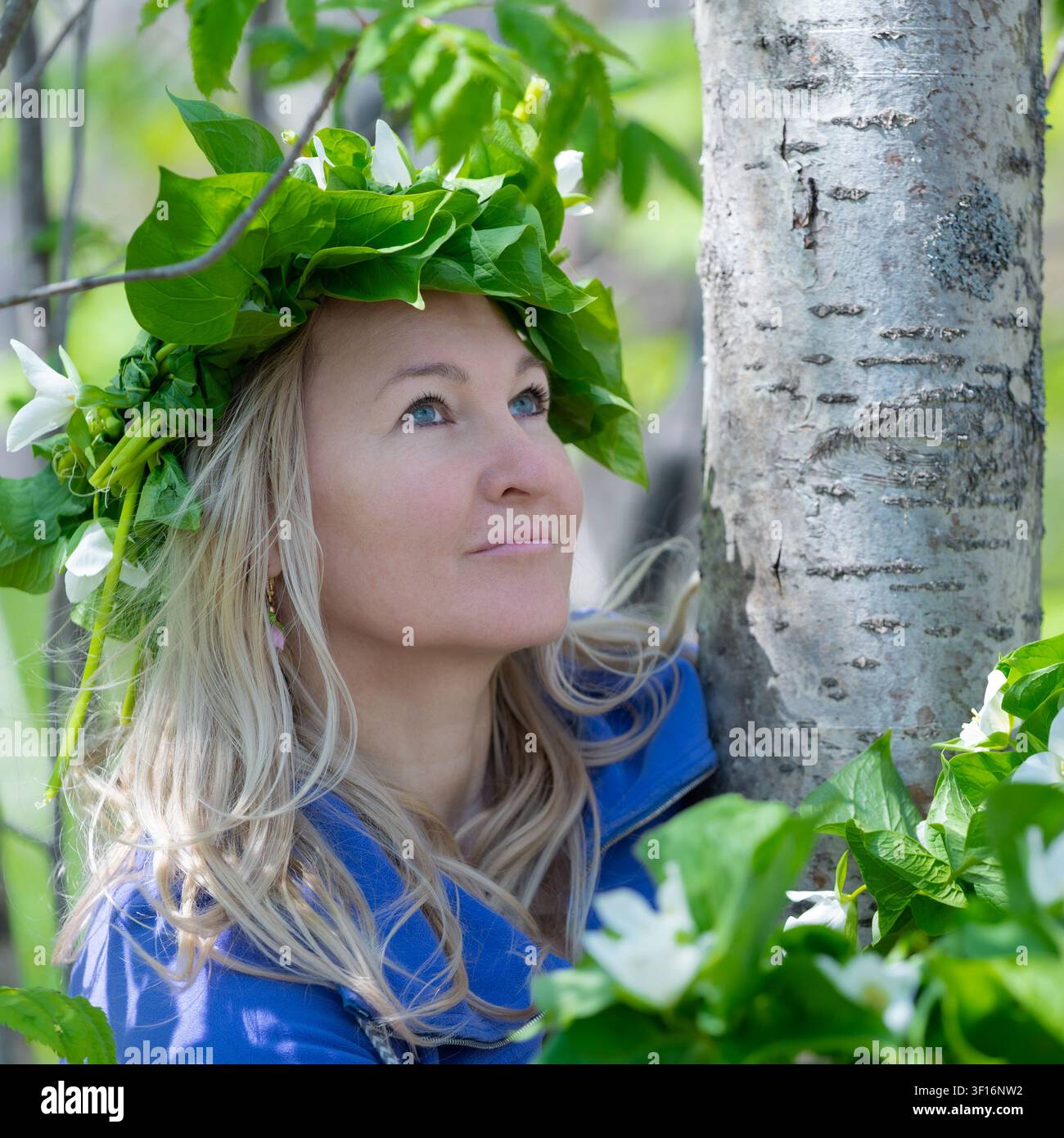 Femme blonde en veste bleue devant un bouleau entouré de fleurs de trillium blanc. Une lumière naturelle douce met en valeur une expression sereine Banque D'Images